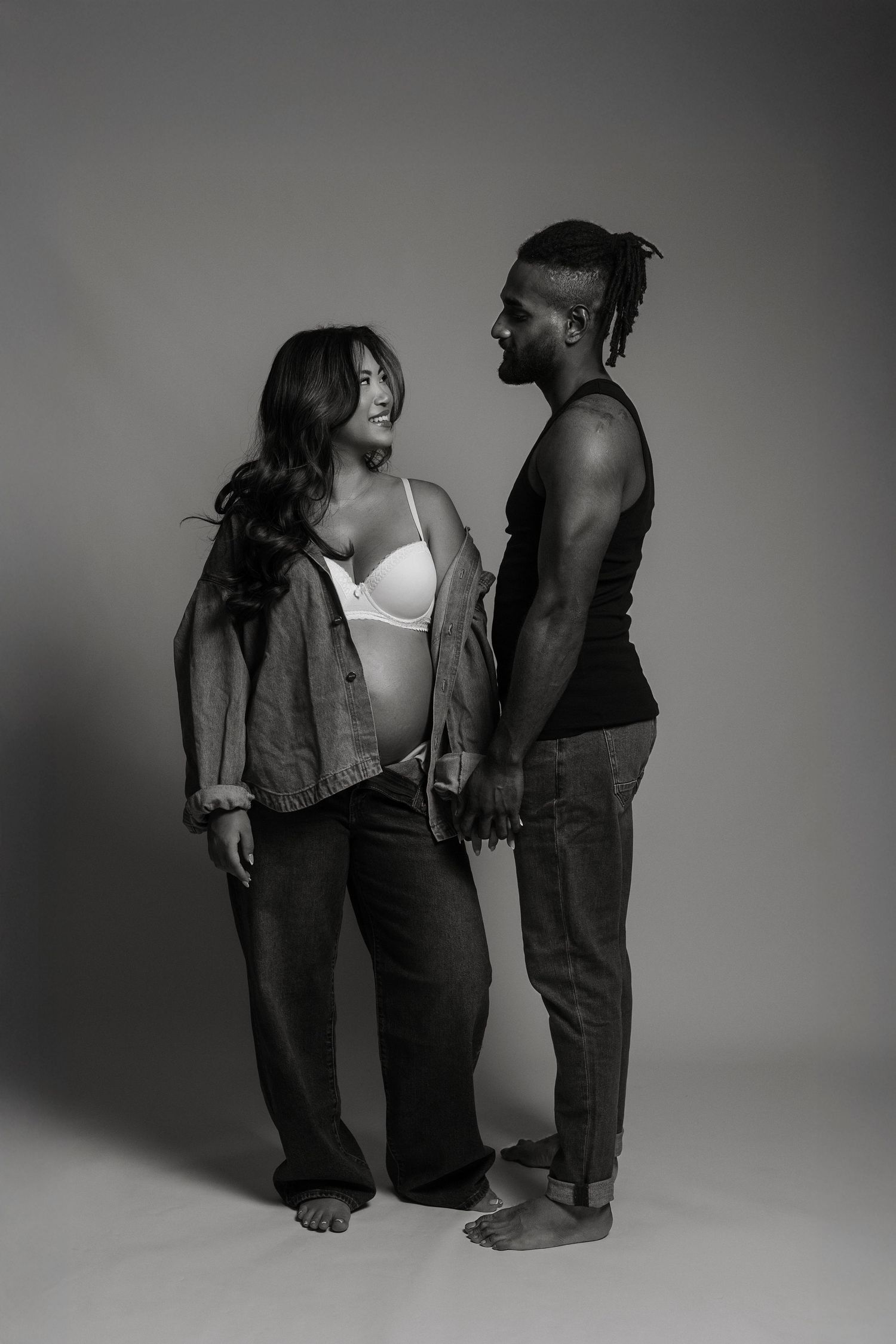 Black and white photo of a pregnant woman and man holding hands, smiling at each other in a studio setting.