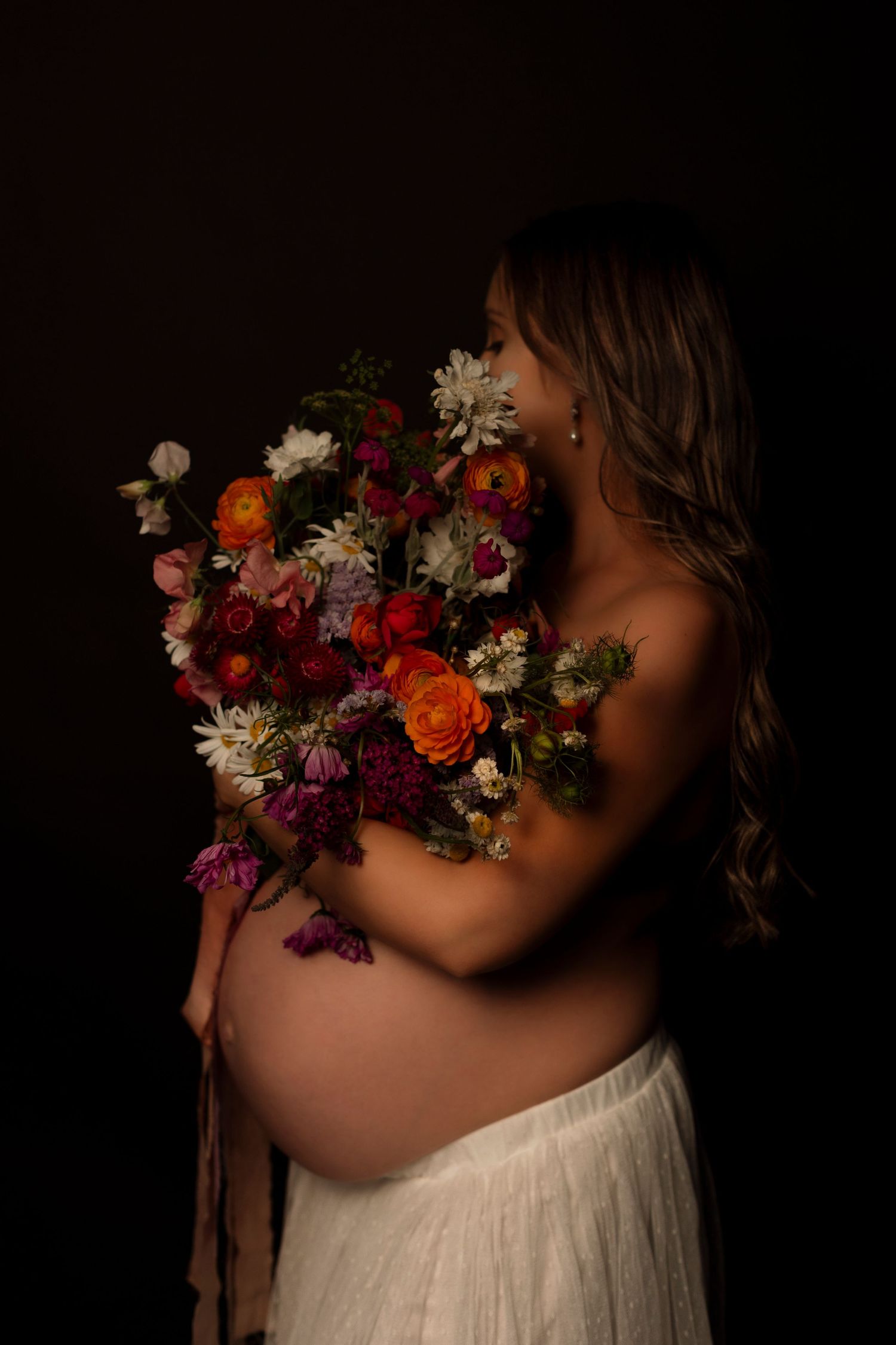 Artistic maternity photo with colorful floral arrangement against dark background.