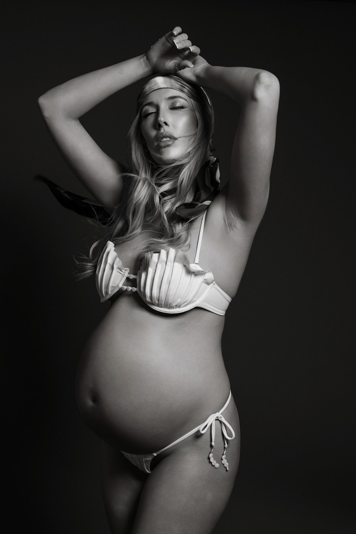 Artistic black and white maternity photo of an expectant mother in lingerie posing against dark background.