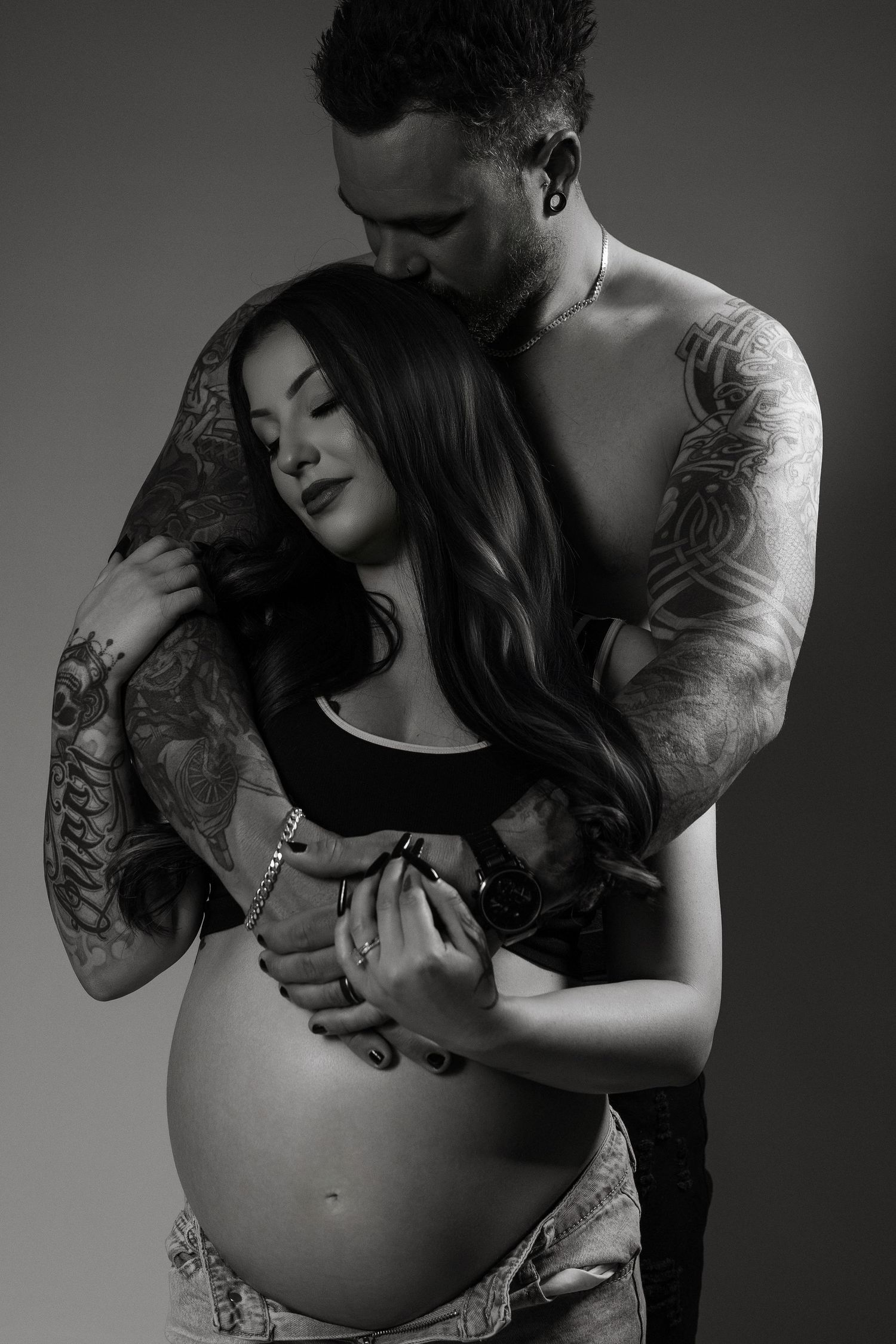 Black and white intimate maternity photo showing a couple embracing, with visible tattoos and a pregnant belly.