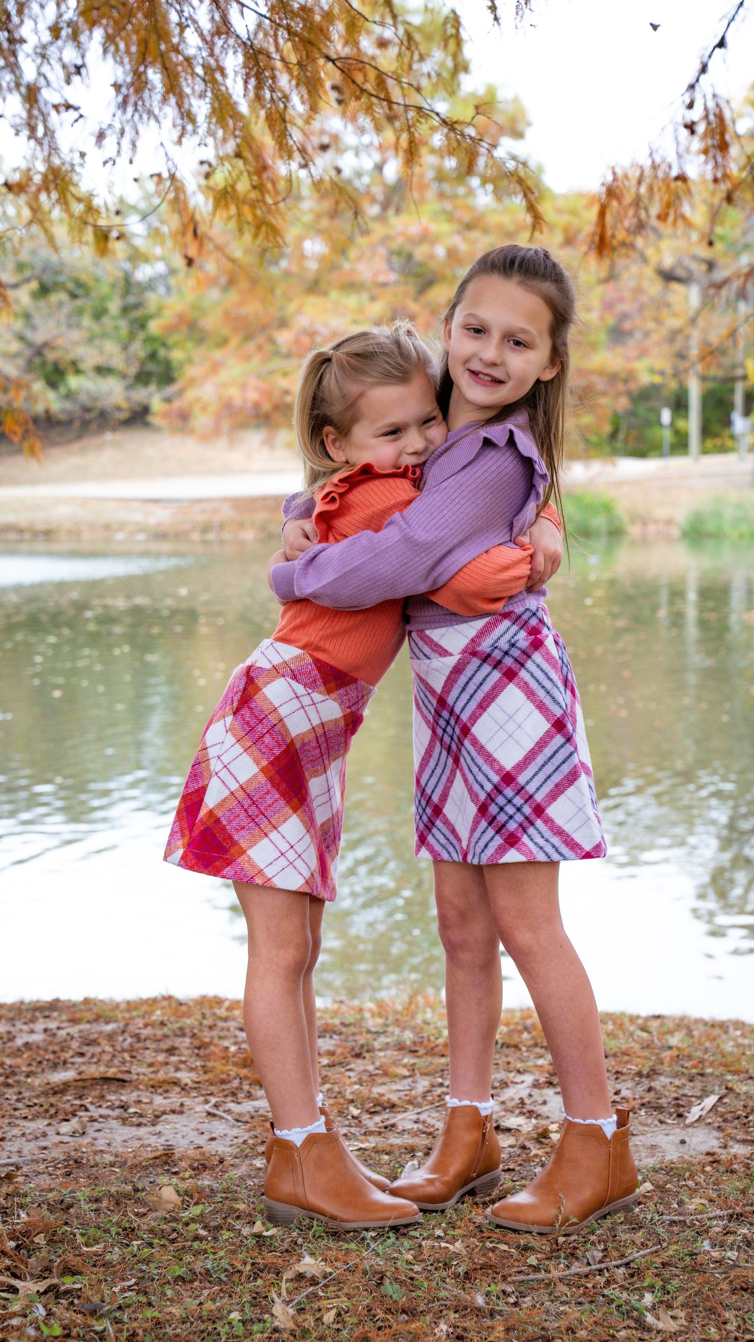 Two children in plaid skirts hugging by a lake on an autumn day with orange foliage and reflective water in the background.