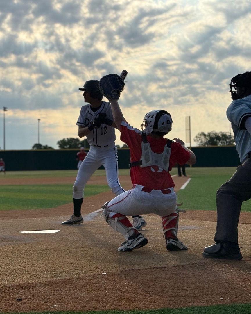 Baseball catcher in red gear signals pitch at home plate during a youth game under dramatic cloudy skies.