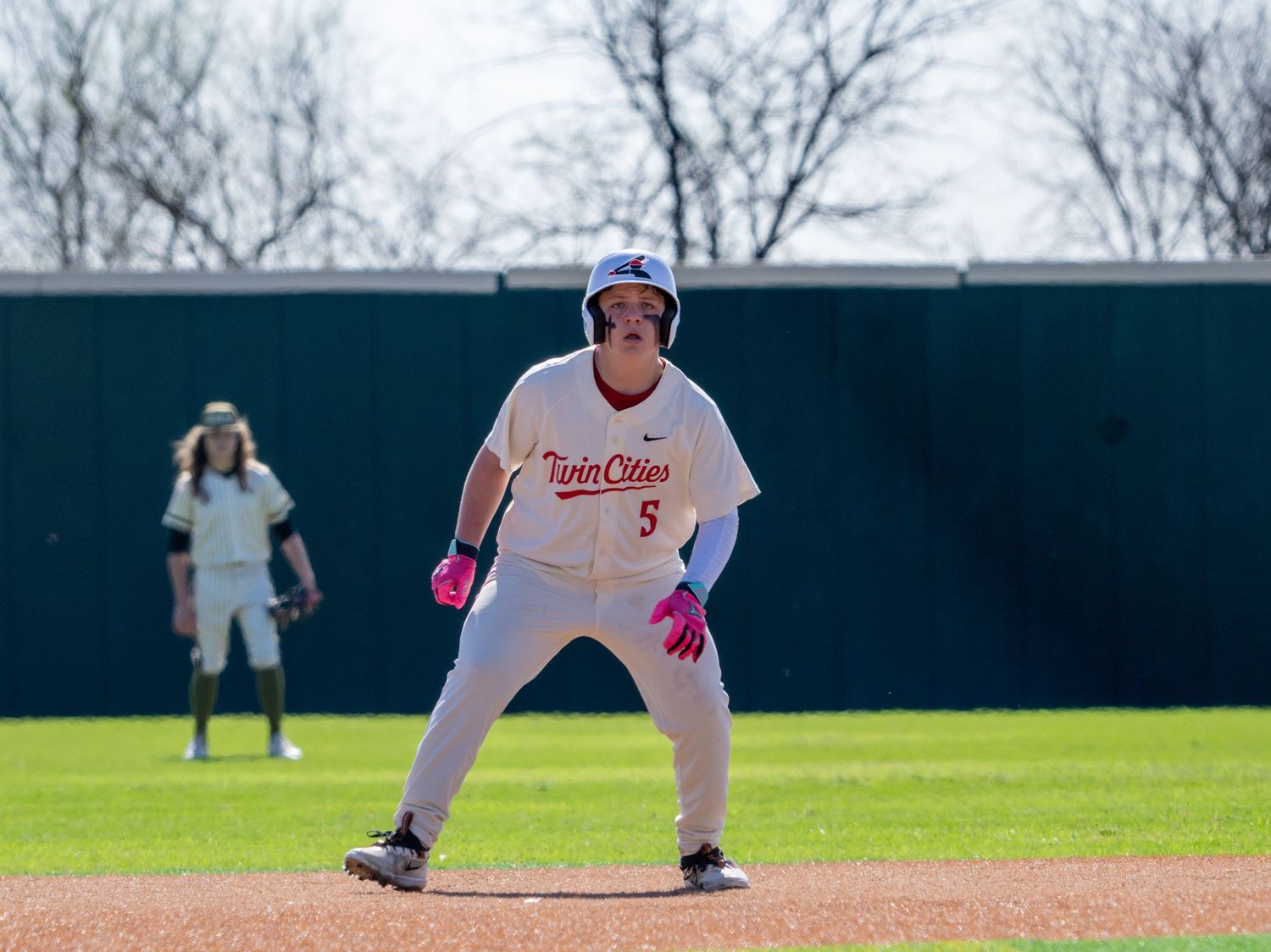 Baseball player wearing number 5 Twin Cities jersey takes a lead off base during a college baseball game.