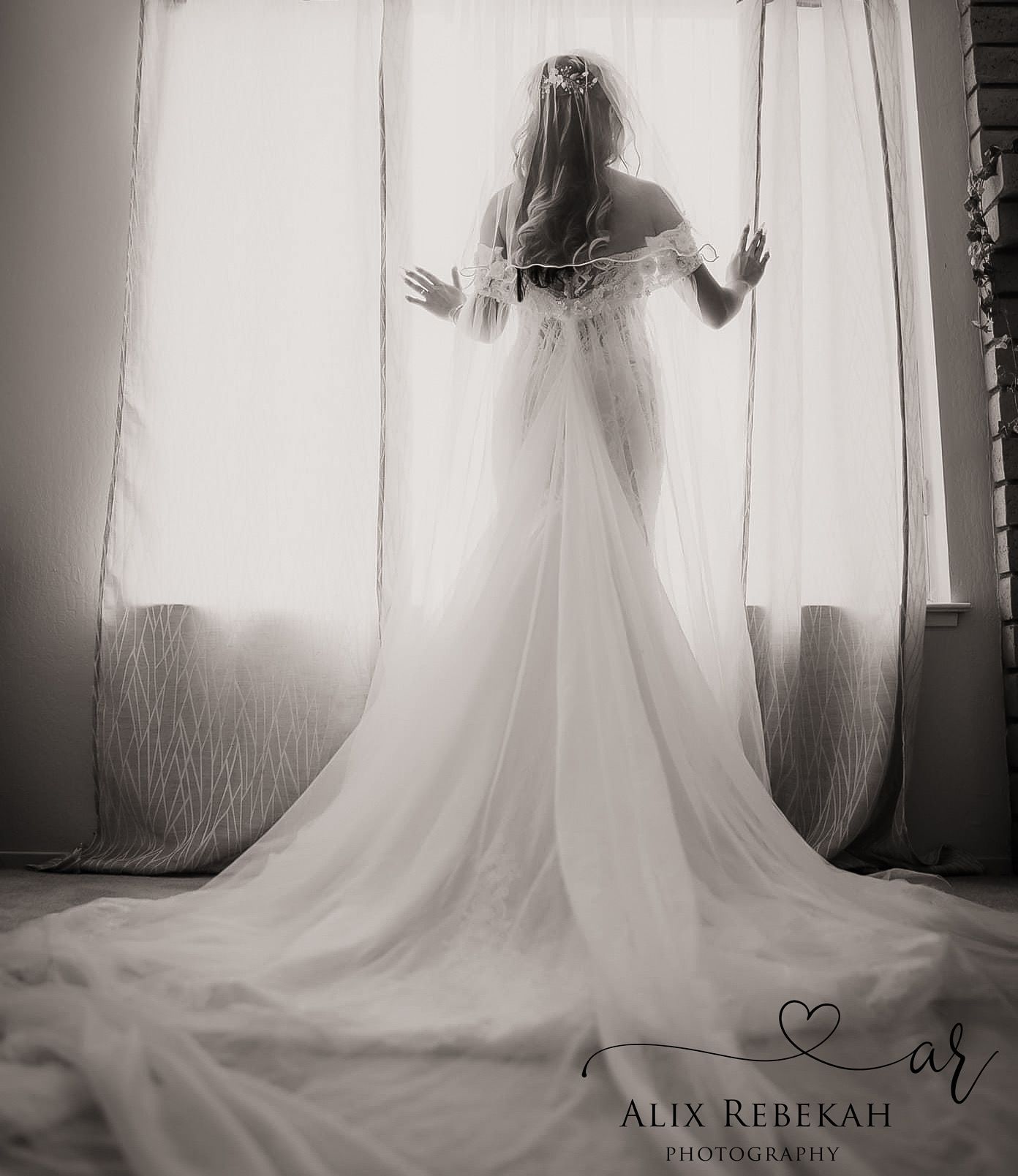 Silhouette of bride in flowing wedding gown standing against backlit curtains in black and white.