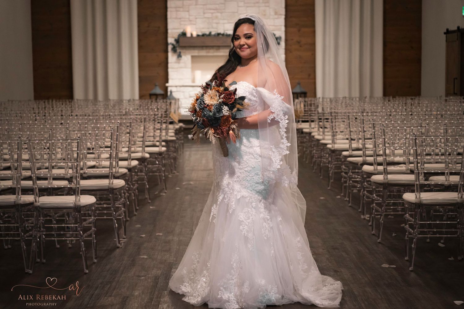 A bride in a lace mermaid wedding dress stands in a church aisle lined with white chairs.