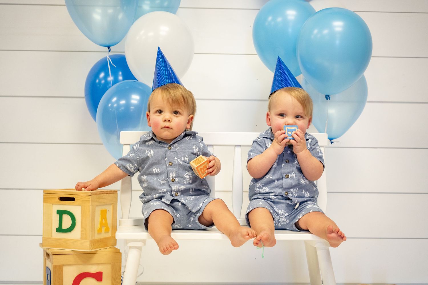 Twin babies in matching outfits pose with alphabet blocks and blue balloons.