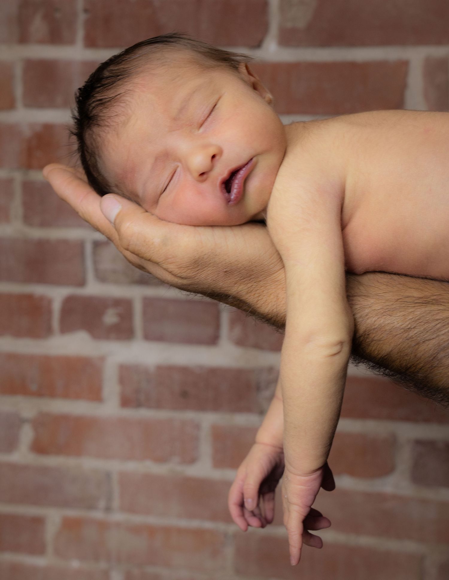 Sleeping newborn cradled in hands against rustic brick wall backdrop.