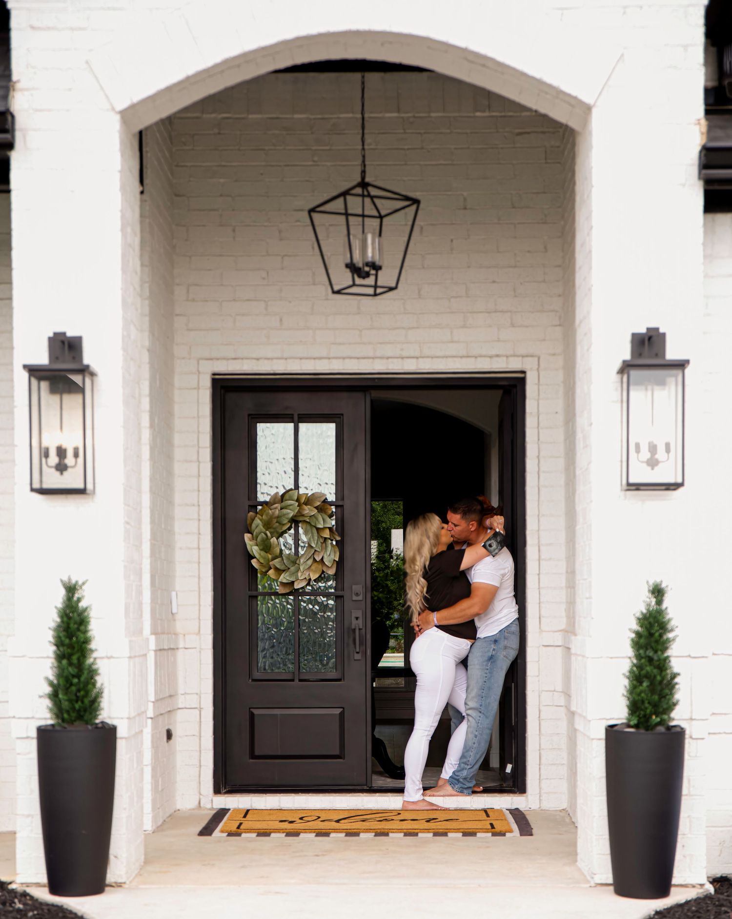 Elegant white brick entryway with black door, lantern lights and potted plants.