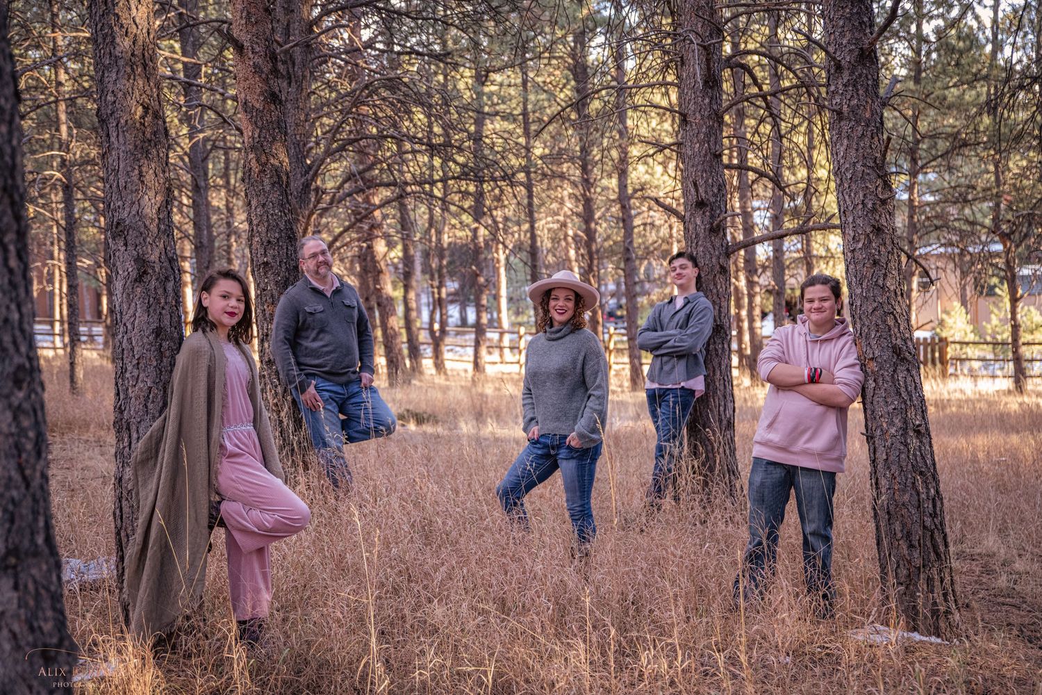 A group poses casually among tall pine trees in a rustic forest setting during golden hour.