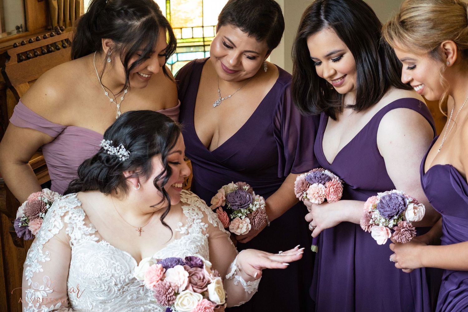 Bridal party in purple dresses gathered together looking at floral bouquets.