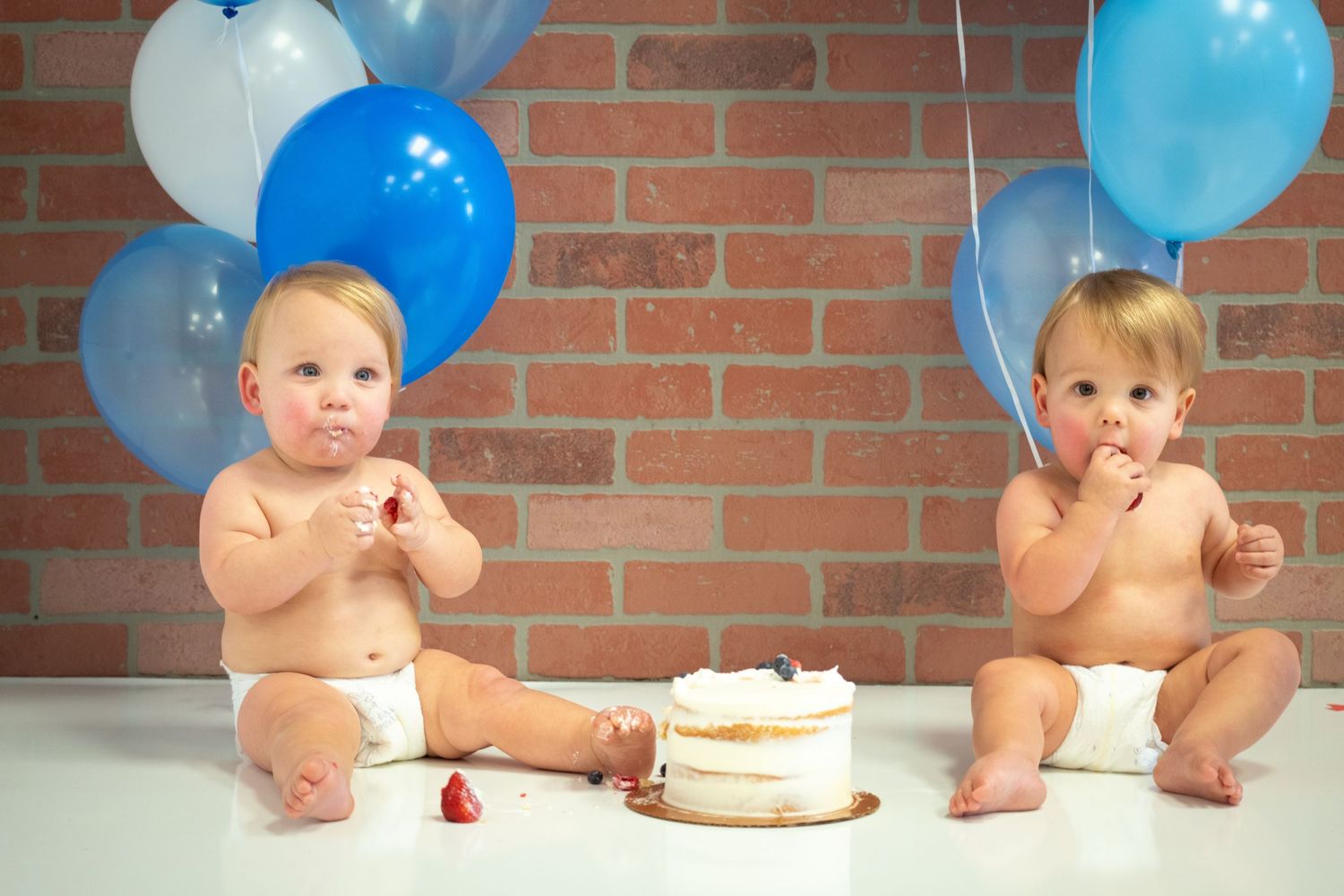 Twin babies in diapers sitting by birthday cake with blue balloons and brick background.