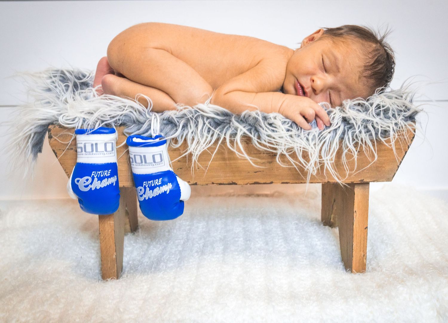 A sleeping baby lying on a wooden bench with blue boxing gloves in a styled newborn photograph.