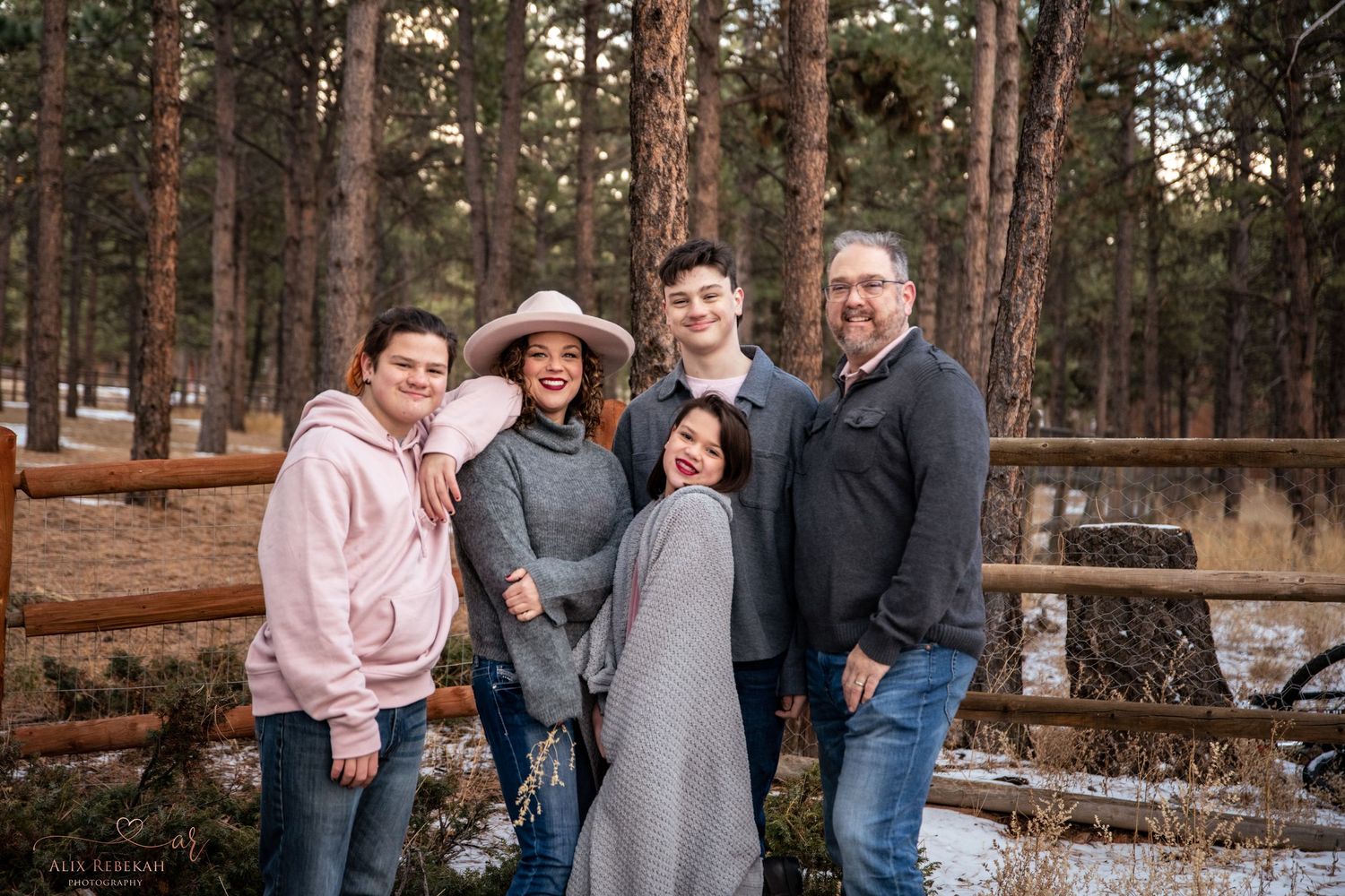 Winter family portrait of group in casual wear standing by rustic wooden fence in pine forest.