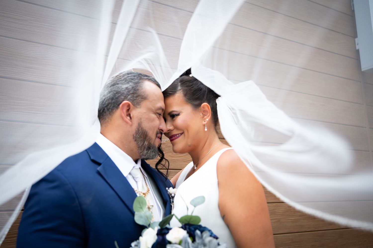 Tender moment captured under flowing white veil with blue and white wedding bouquet.