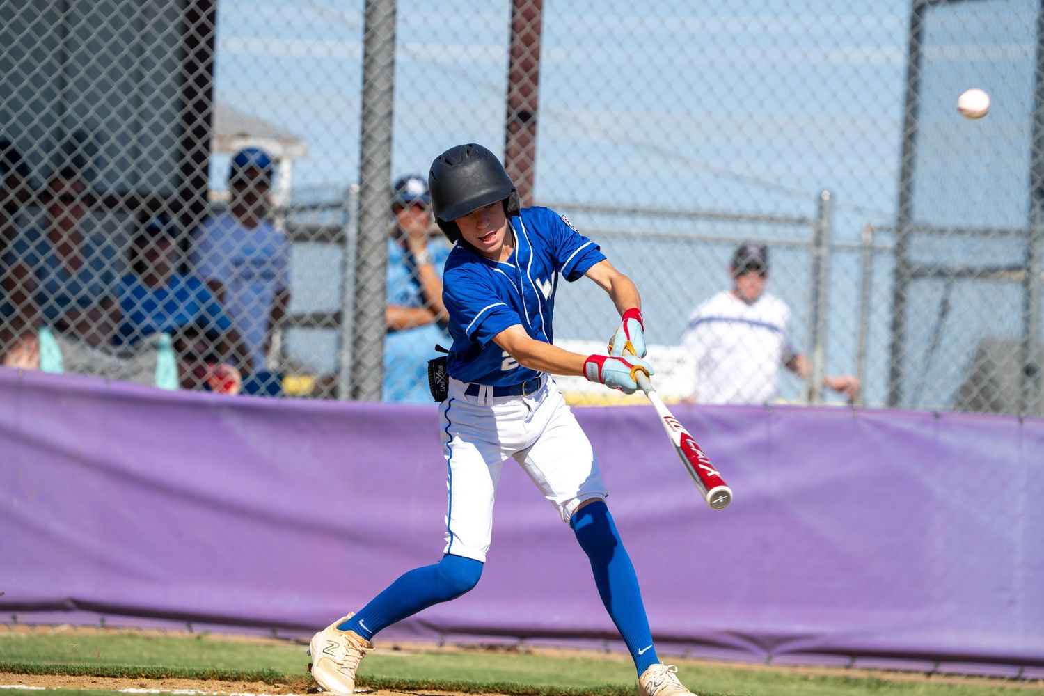 Baseball player in blue jersey and white pants swinging a bat during a game with fenced background and purple banner.