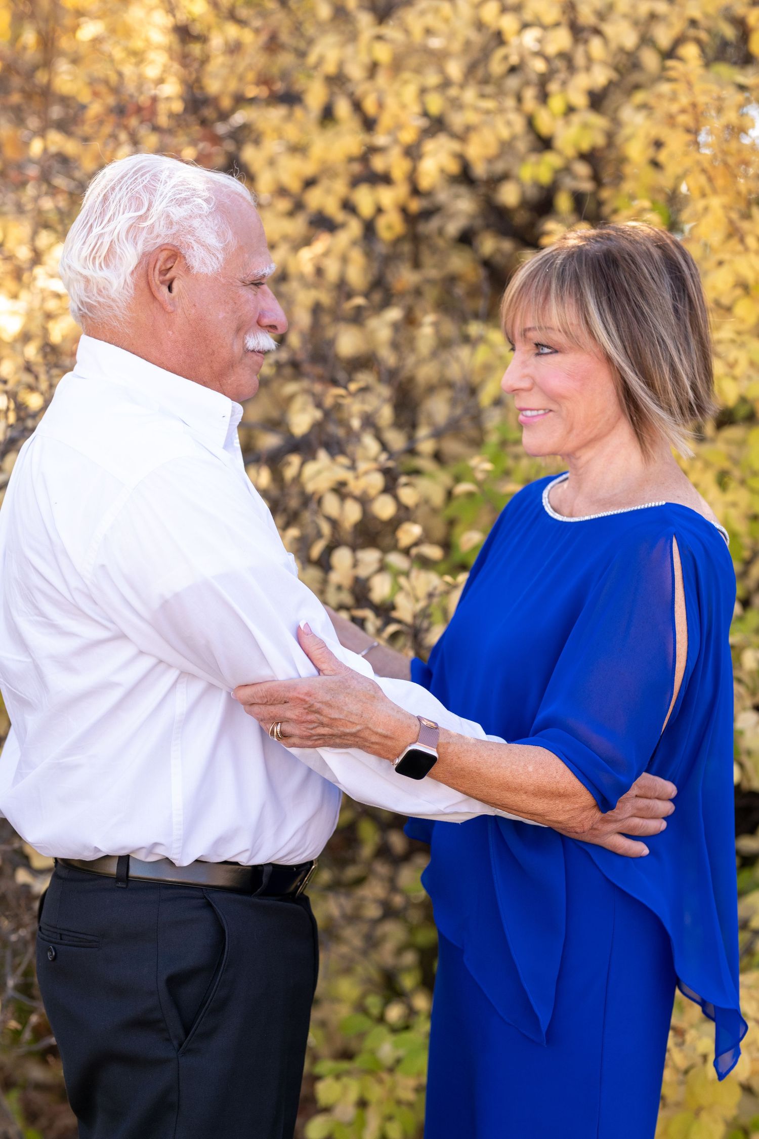 Couple wearing white and blue outfits embrace while looking at each other against autumn foliage background.