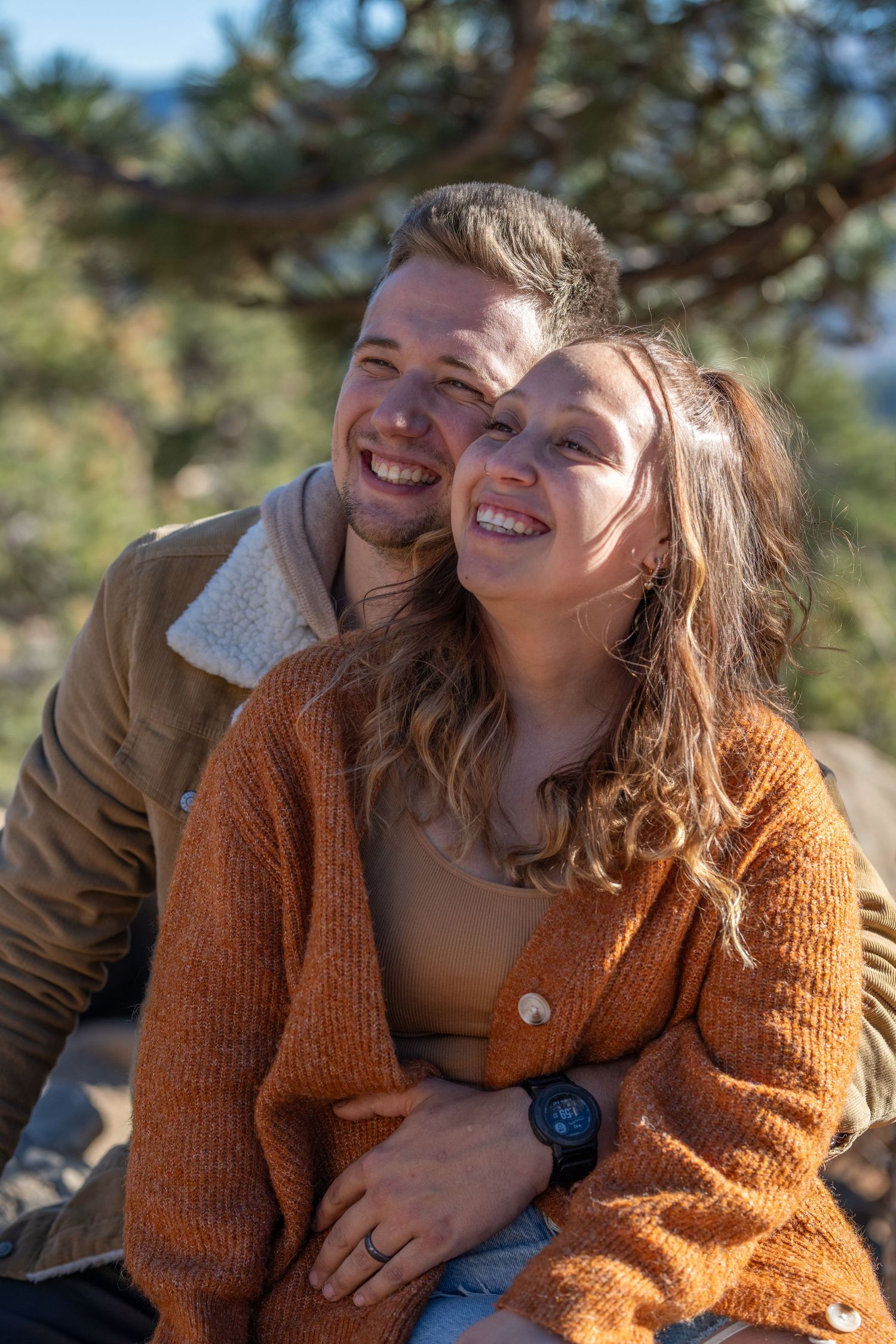 A couple shares a candid laughing moment outdoors with pine trees in the background.