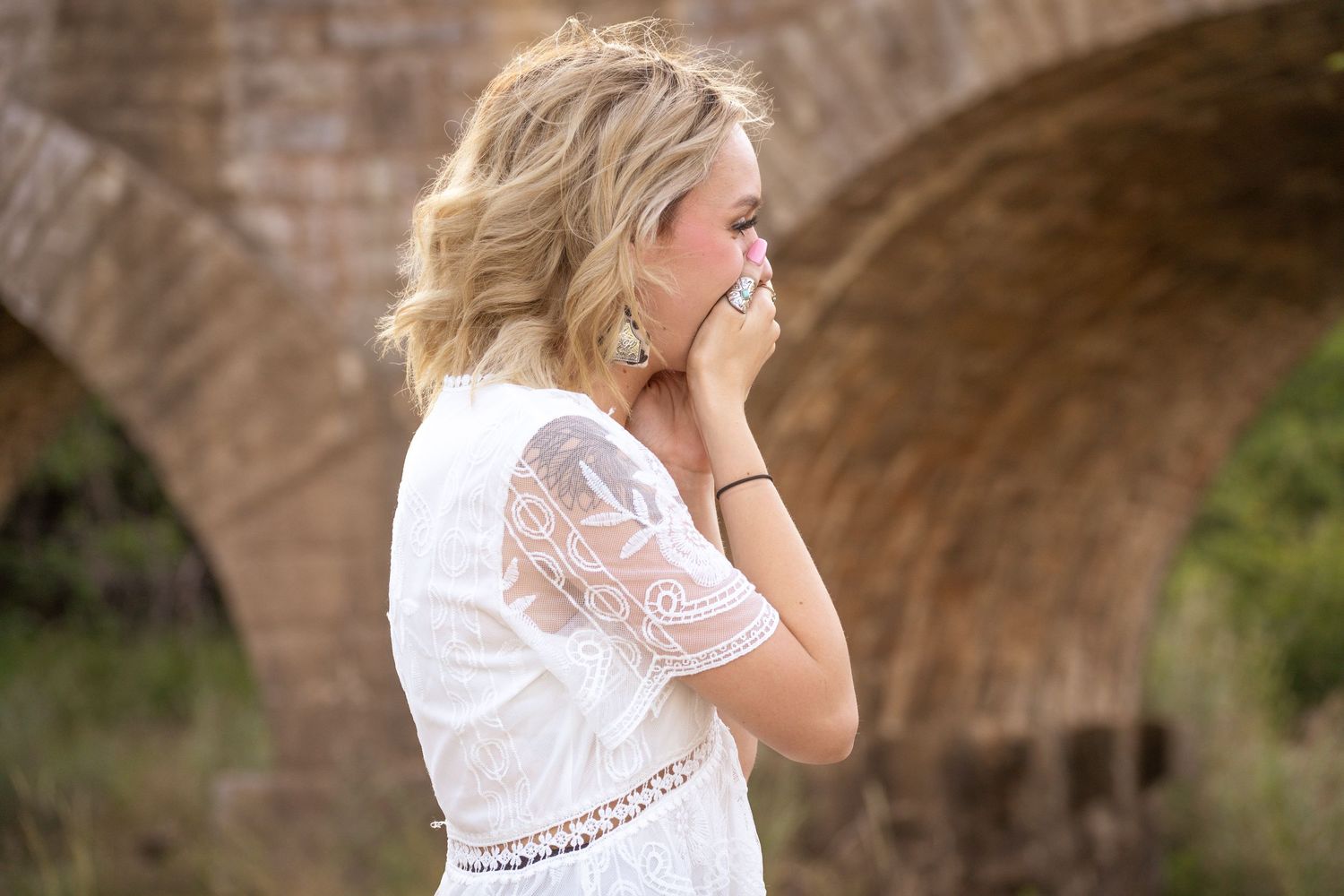 Person in white lace dress stands near stone archway with hand covering mouth in emotional moment.