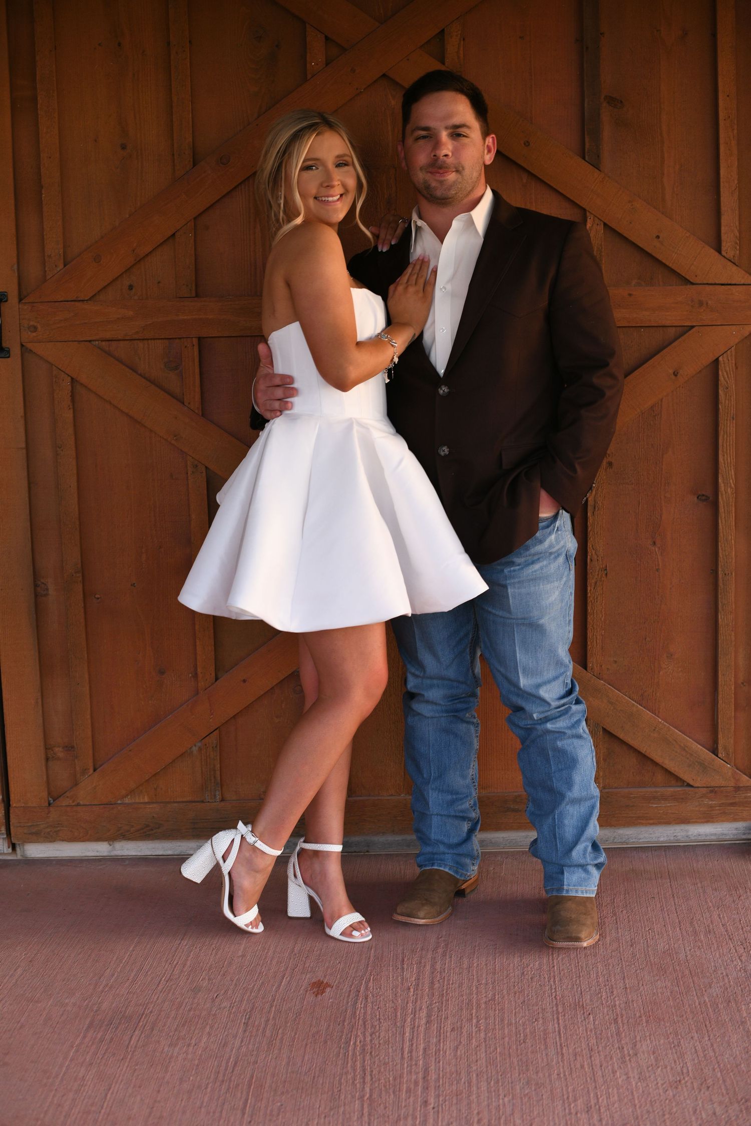 Couple poses in front of rustic wooden barn door, with white cocktail dress and casual western attire.