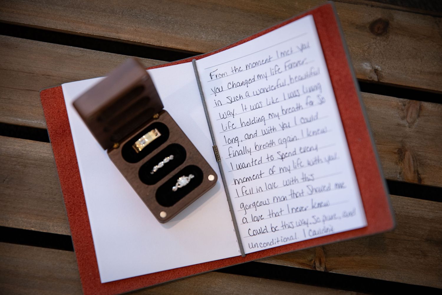 Wedding rings and handwritten vows in a red journal on wooden surface.