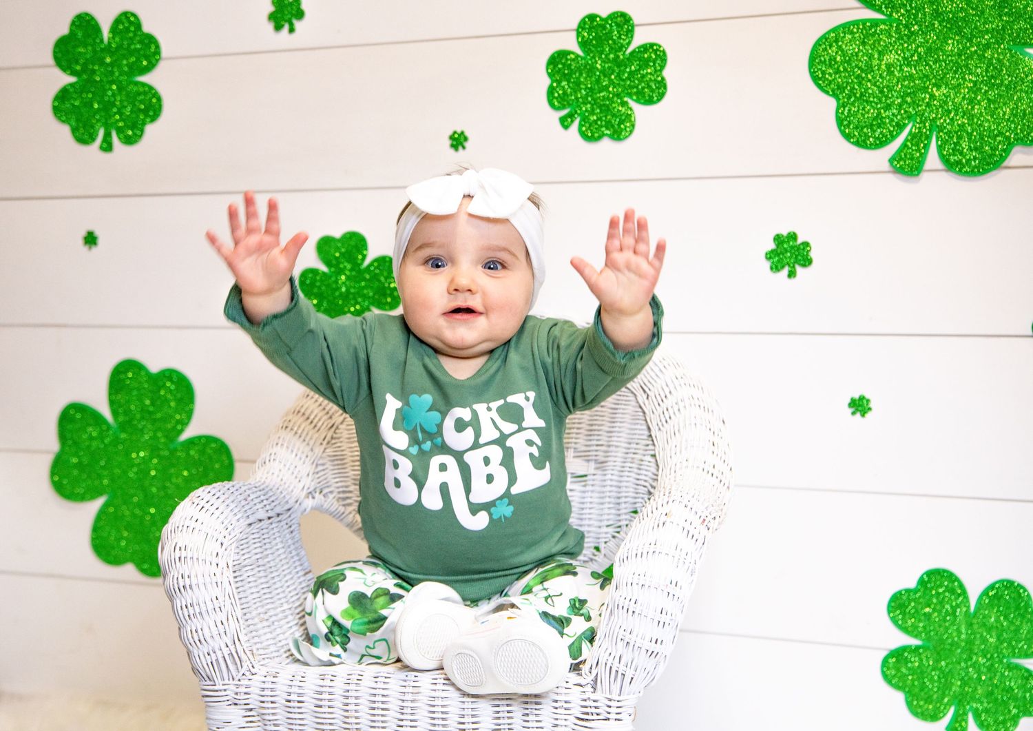 A baby in festive green attire raises hands while sitting in wicker chair with shamrock decorations.