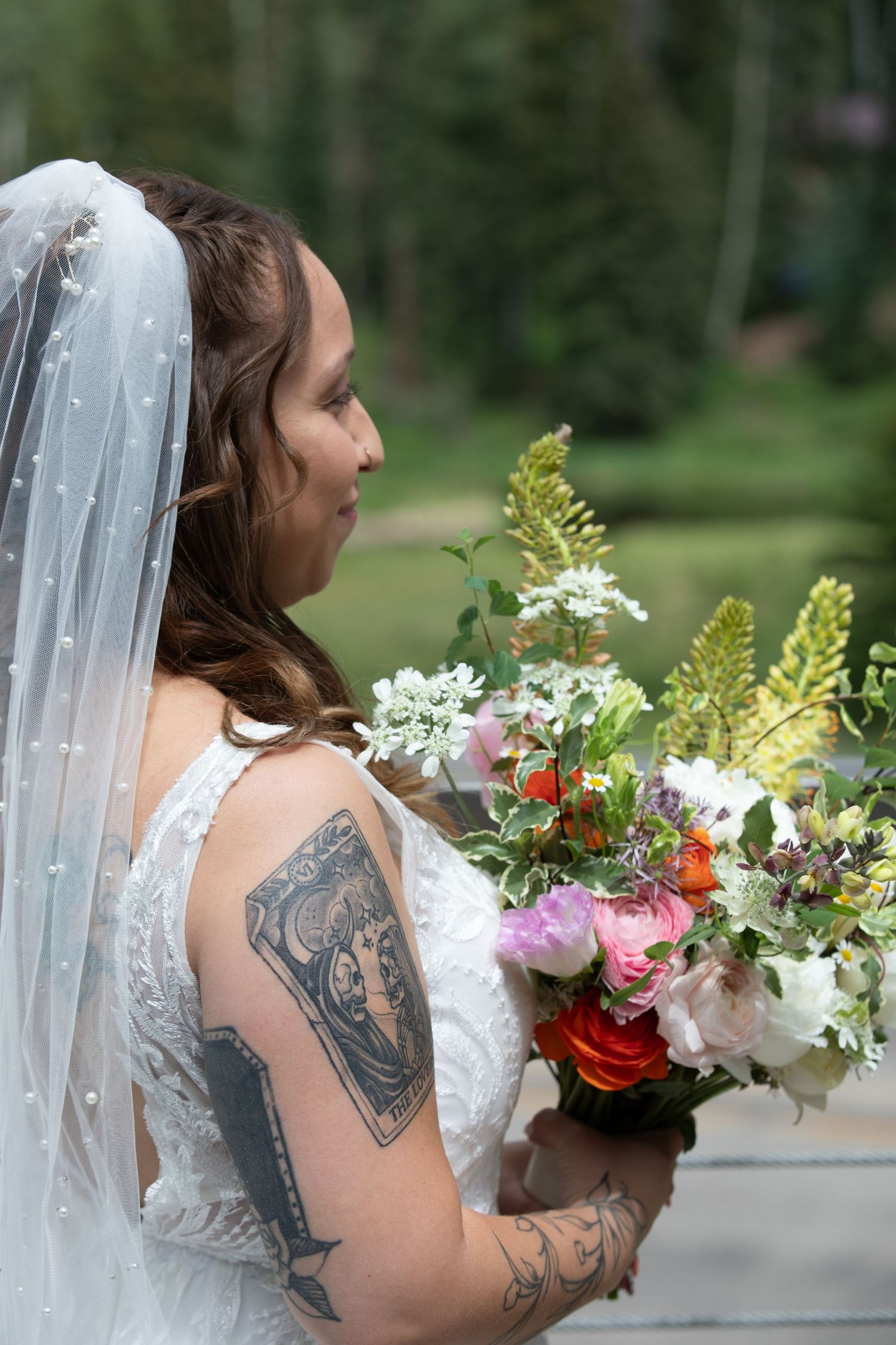 A bride in white dress with tattoos holding a colorful bouquet outdoors.