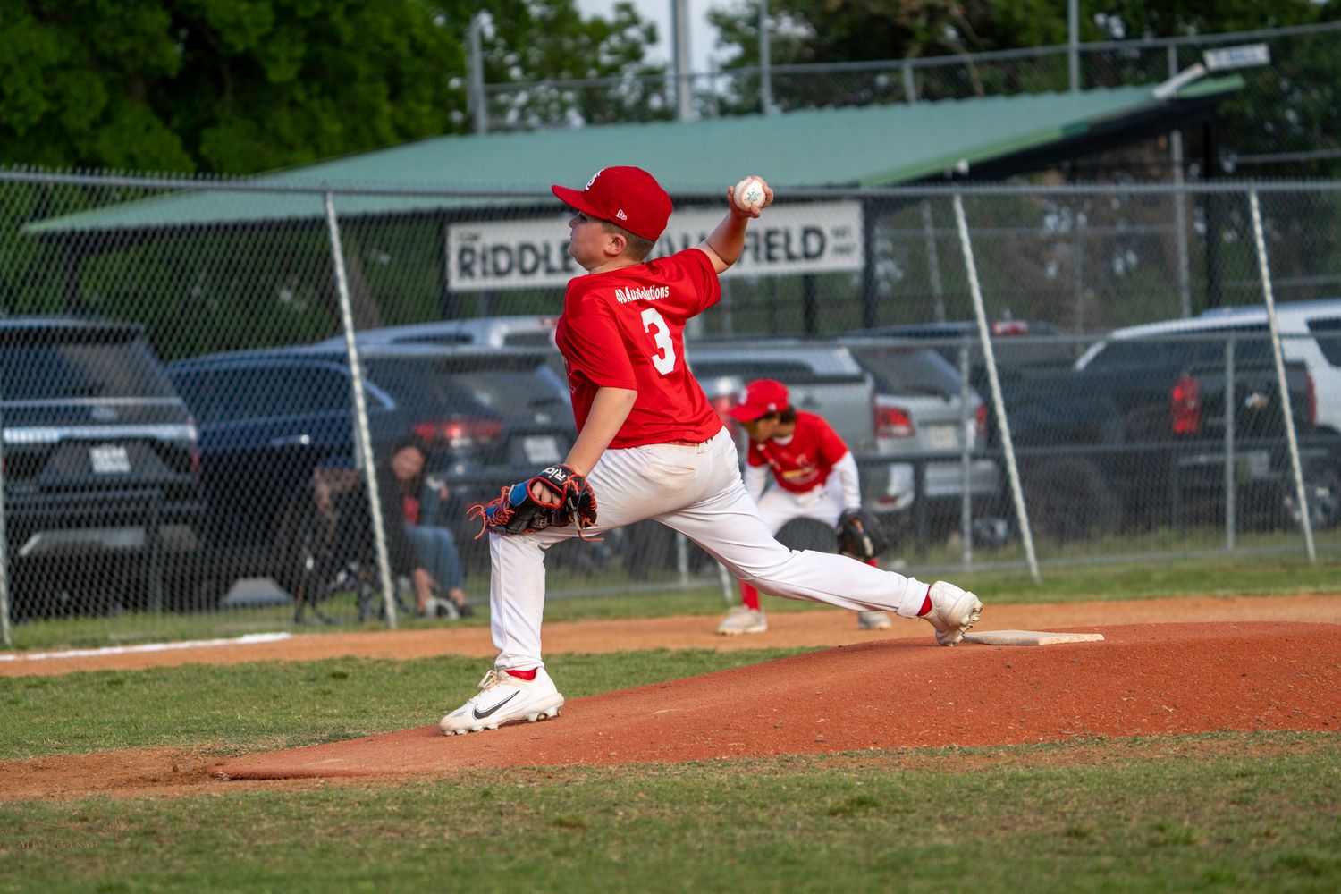 Baseball pitcher in red jersey mid-delivery throwing from the mound during a game on outdoor baseball field.