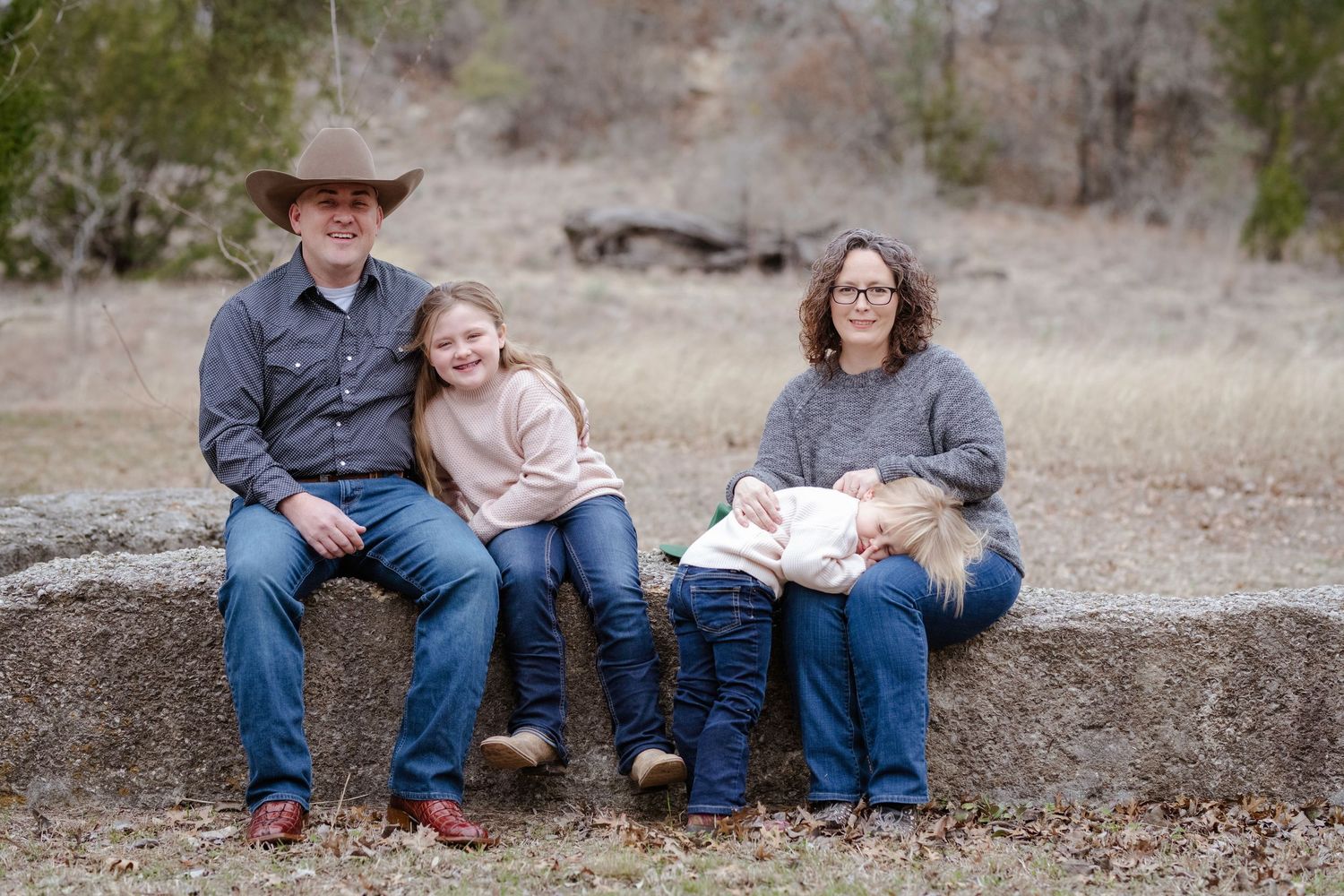 Family of three sits on fallen tree trunk in rustic outdoor setting wearing casual clothes.
