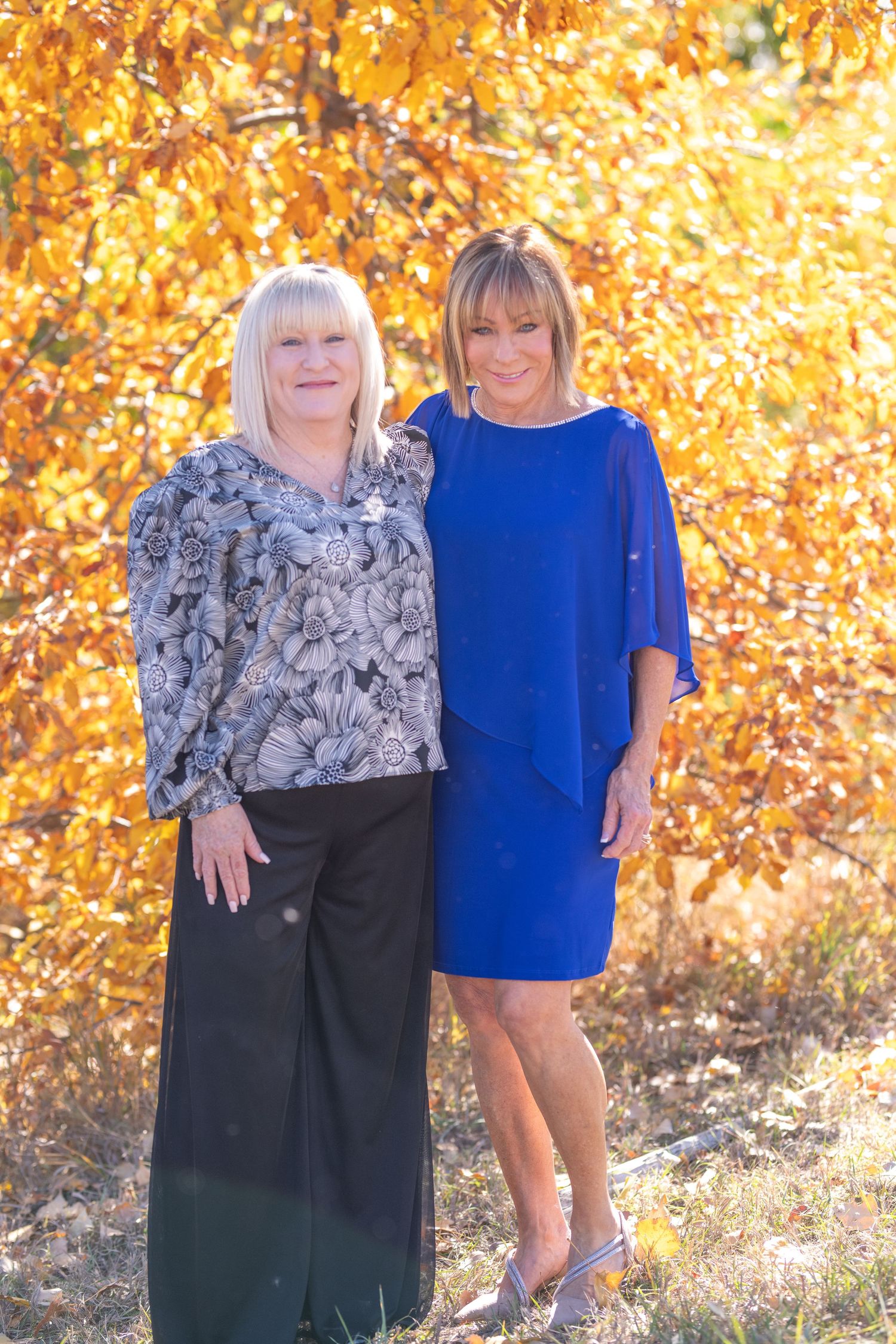 Two people wearing contrasting outfits pose together against a backdrop of vibrant golden autumn foliage.
