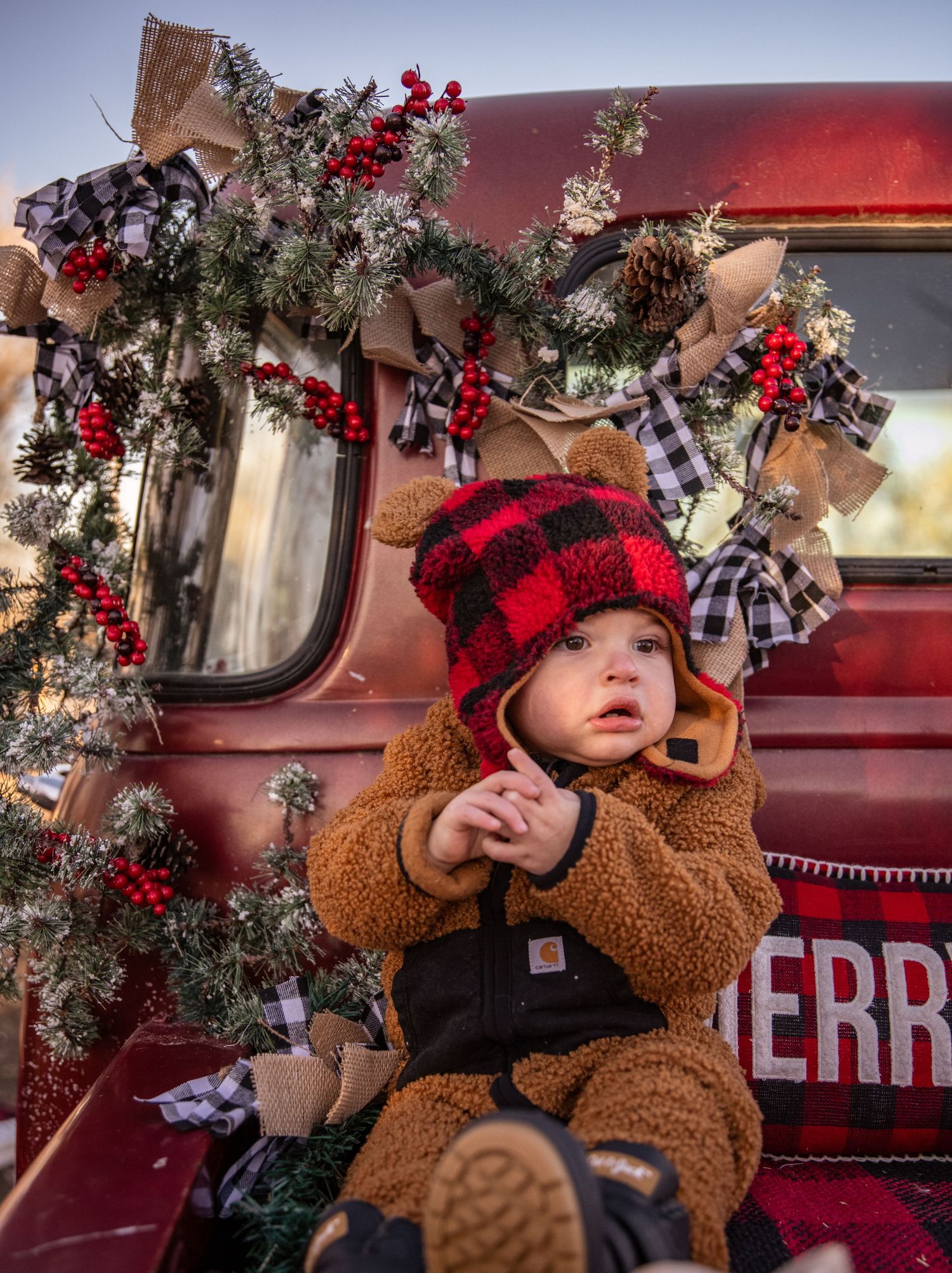 Baby in plaid hat and brown teddy outfit sits in decorated vintage red truck with holiday greenery.