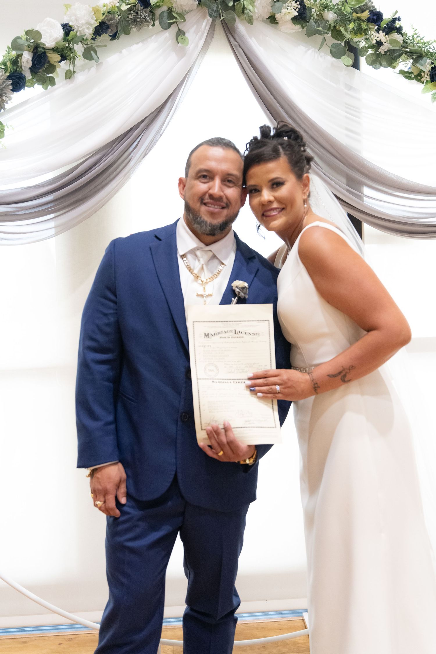 Newlyweds pose with their marriage certificate under an elegant floral arch with white draping.