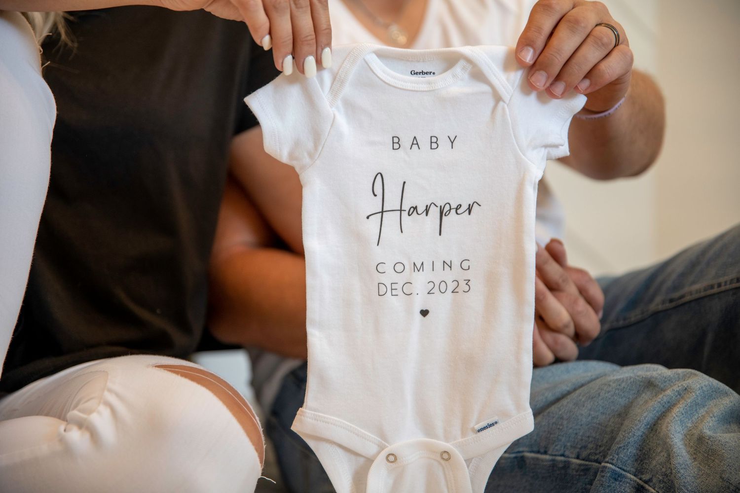 Hands holding up a white baby onesie with text announcing December 2023 arrival.