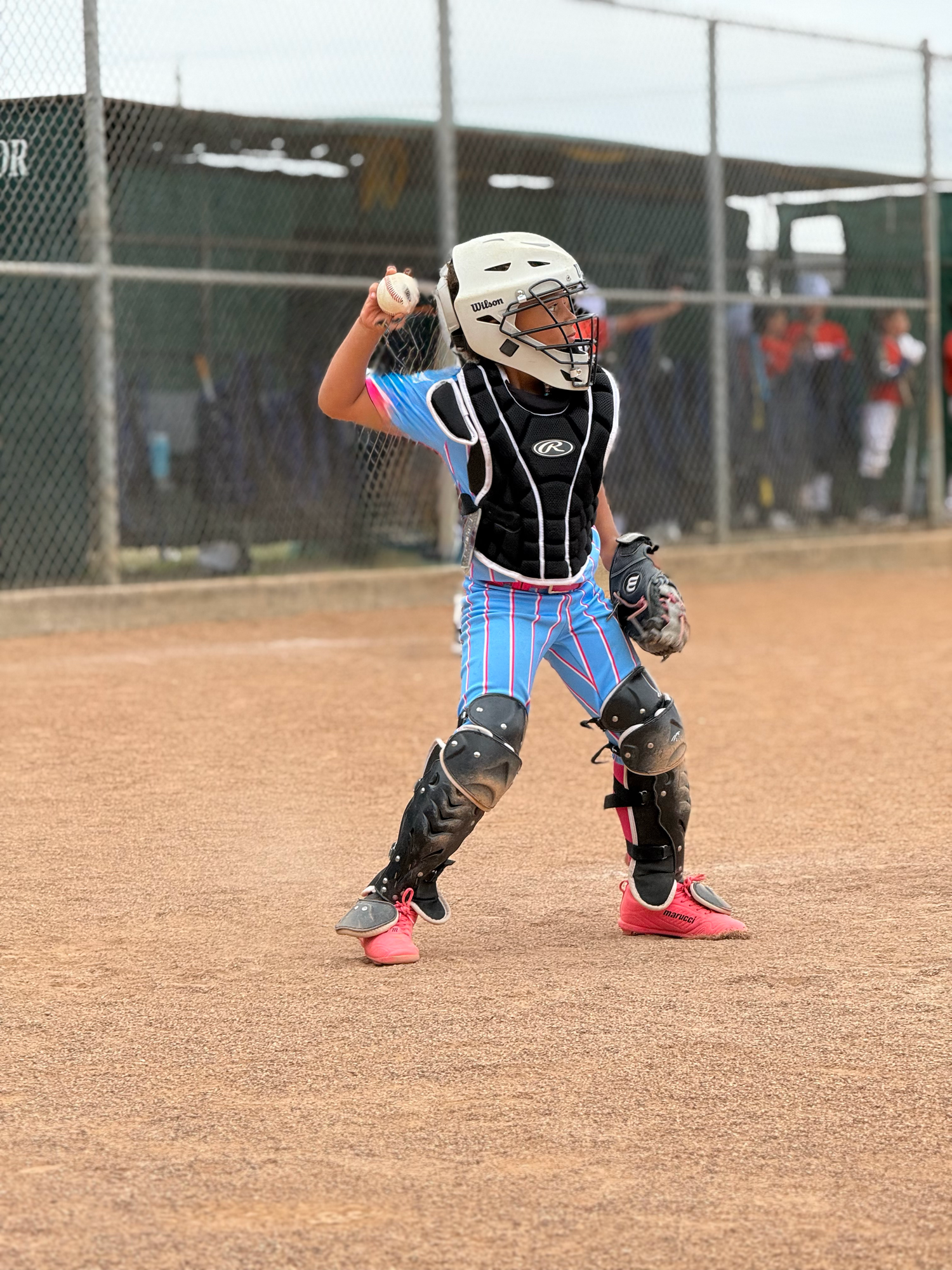 Young baseball catcher in full gear winds up to throw ball on dirt field during youth league game.