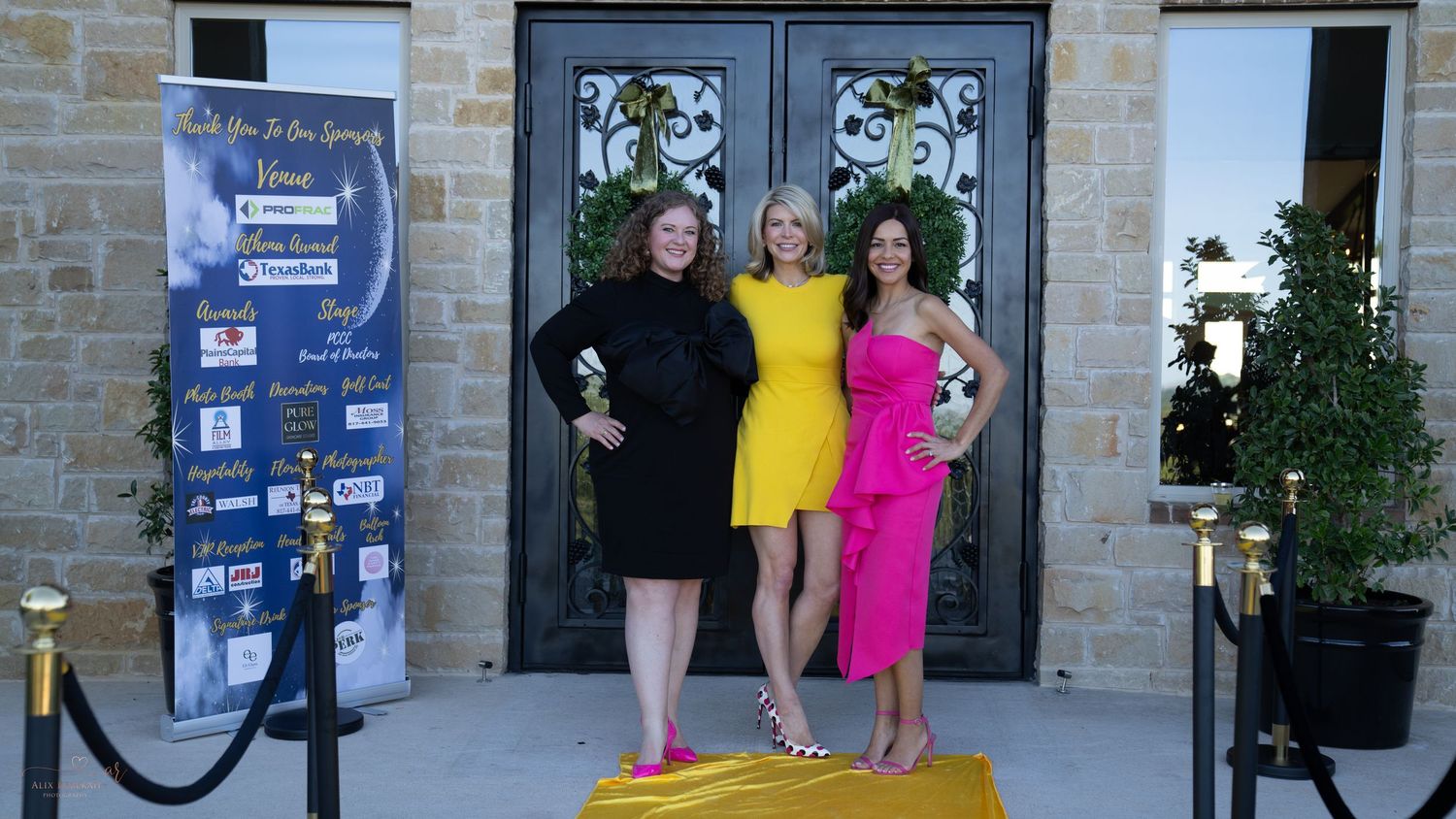 Three people pose together in formal attire on a red carpet at an event entrance.