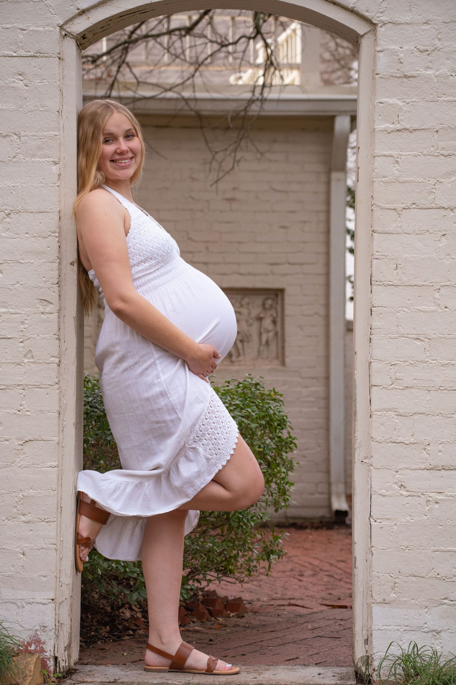 A person in a white lace dress poses in a doorway showing their pregnant belly.