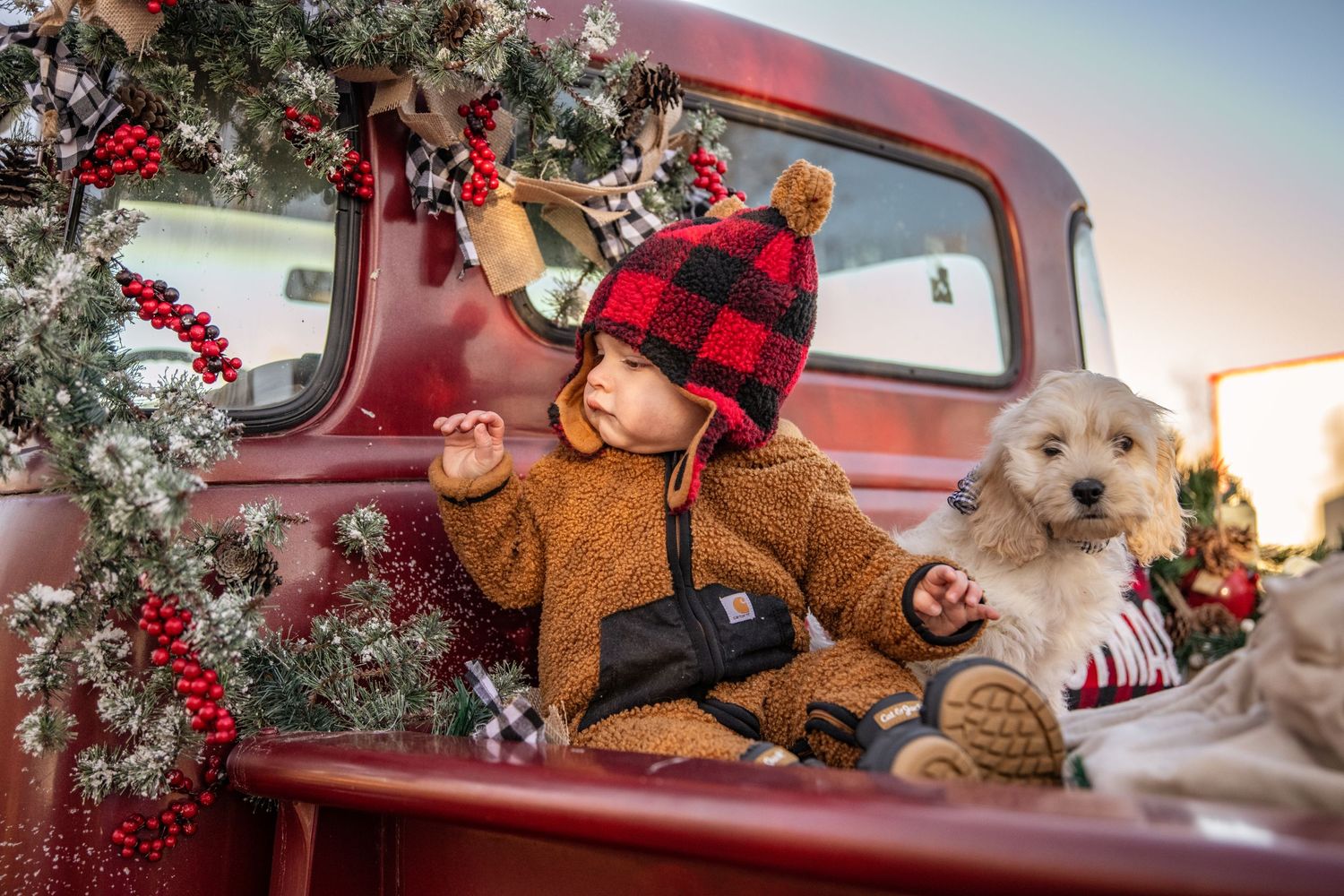Holiday scene with decorated red truck, small child in brown outfit, and fluffy white dog.