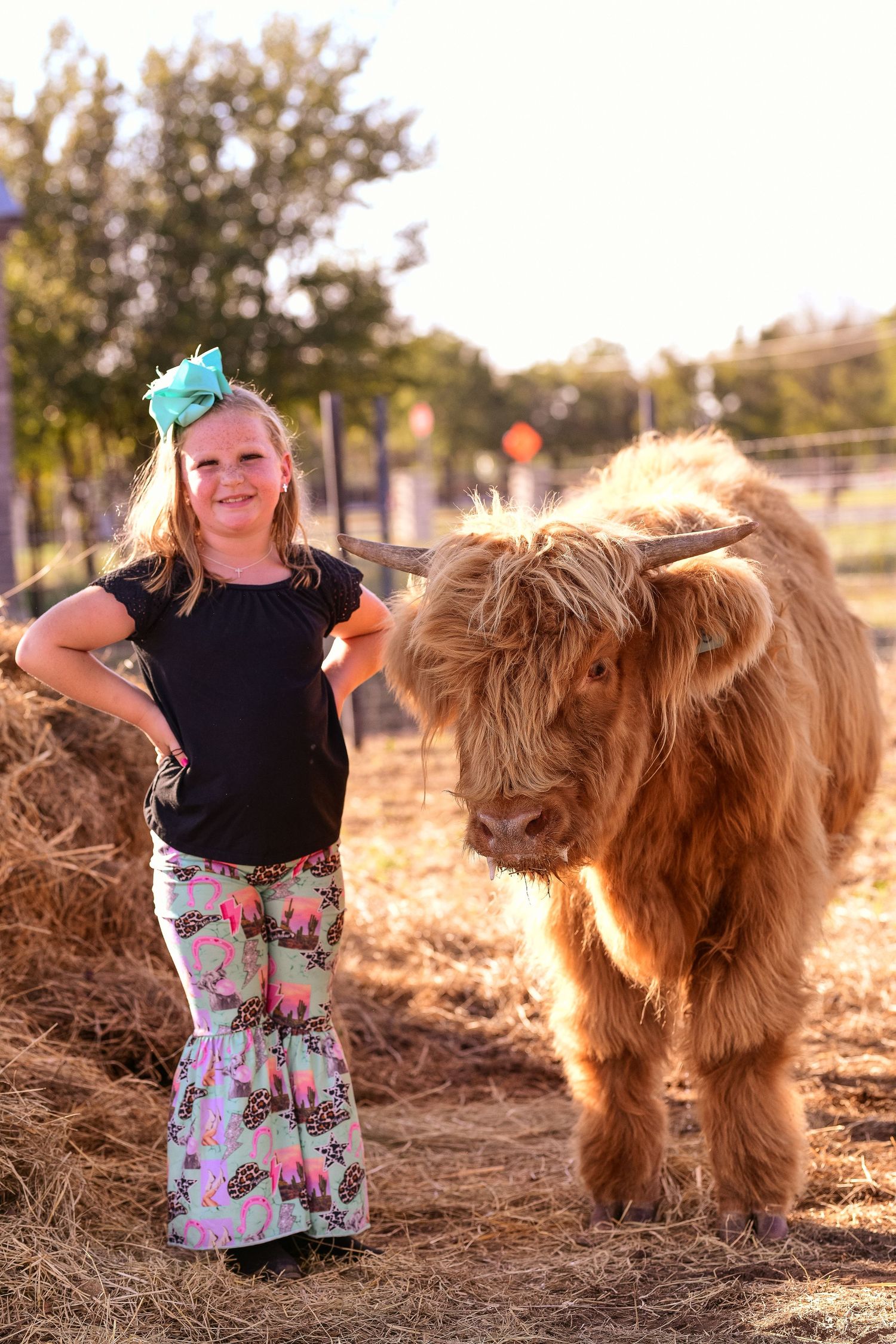 A young person in floral pants poses confidently next to a Highland cow on a farm.