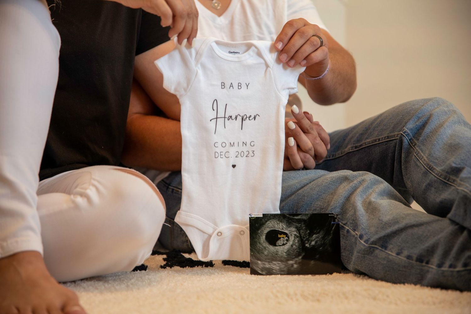 Hands holding white baby onesie with sonogram photo on carpet.