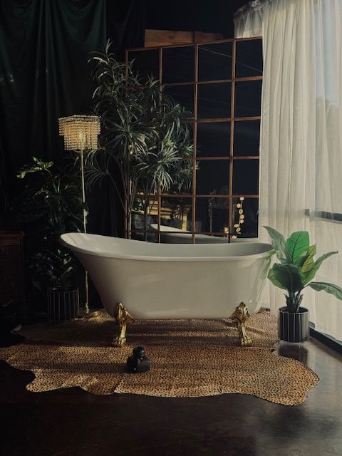 Elegant clawfoot bathtub surrounded by plants and woven rug in a dark moody bathroom setting.