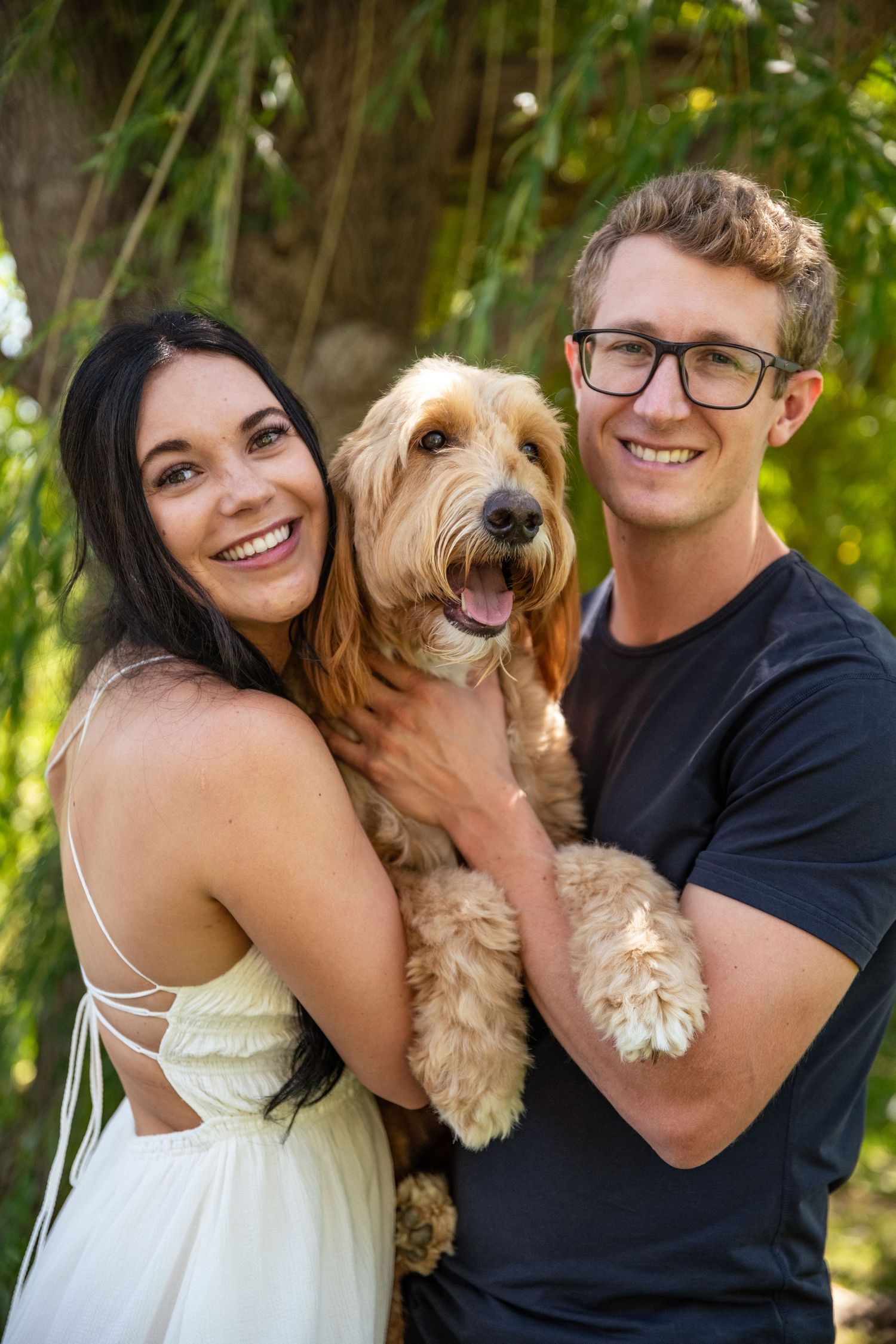Couple holding their happy golden doodle dog while posing under green trees.