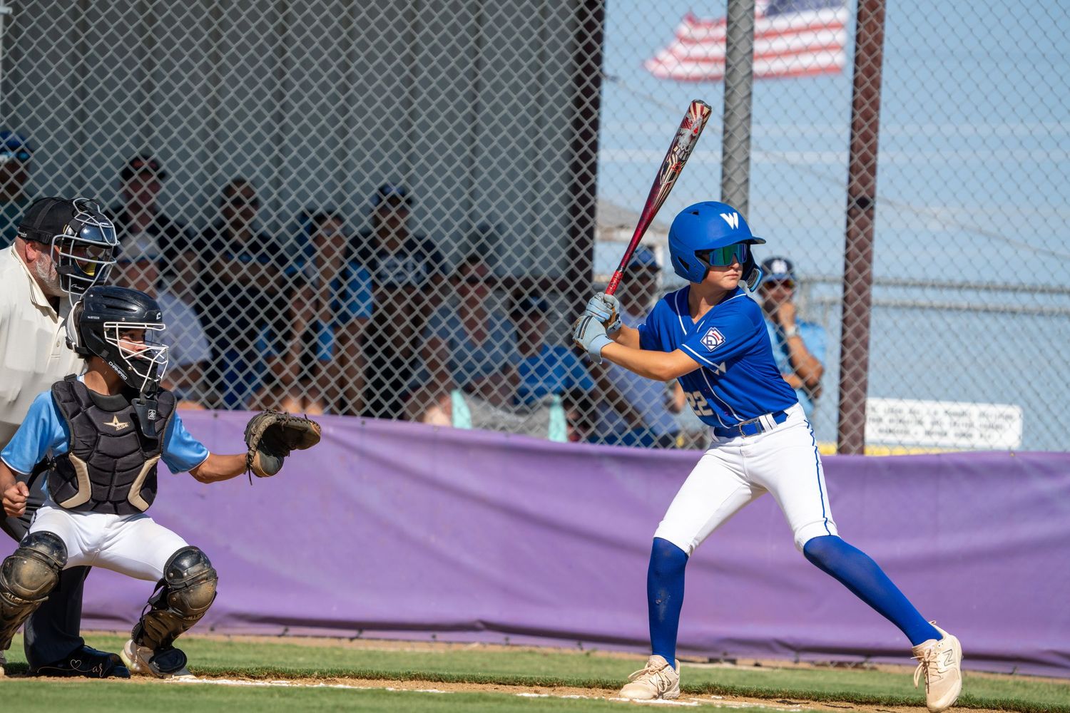 Youth baseball batter in blue uniform at bat while catcher crouches behind home plate during game with American flag visible.