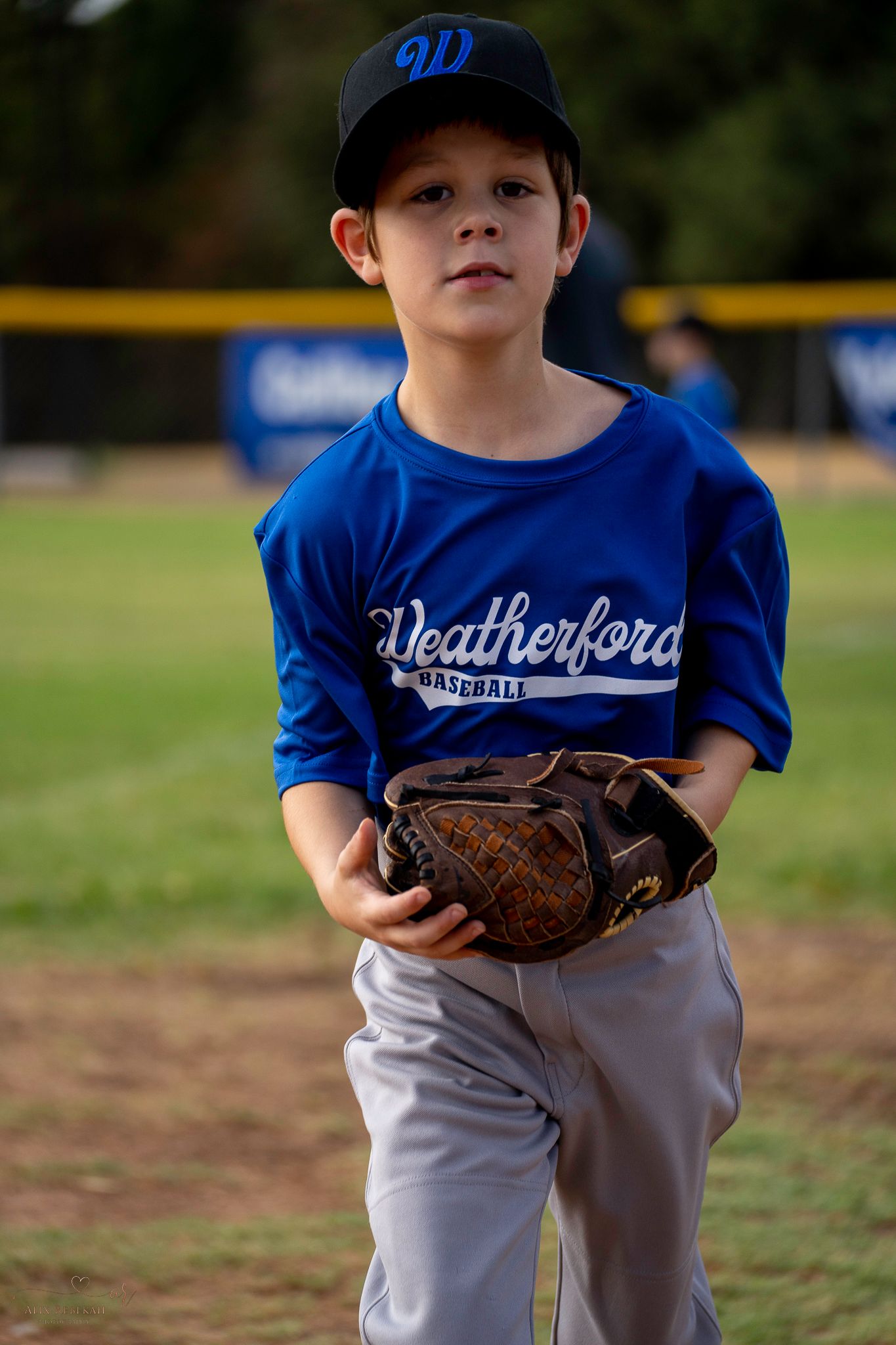Young baseball player in blue Weatherford uniform holds leather mitt on baseball field during game.