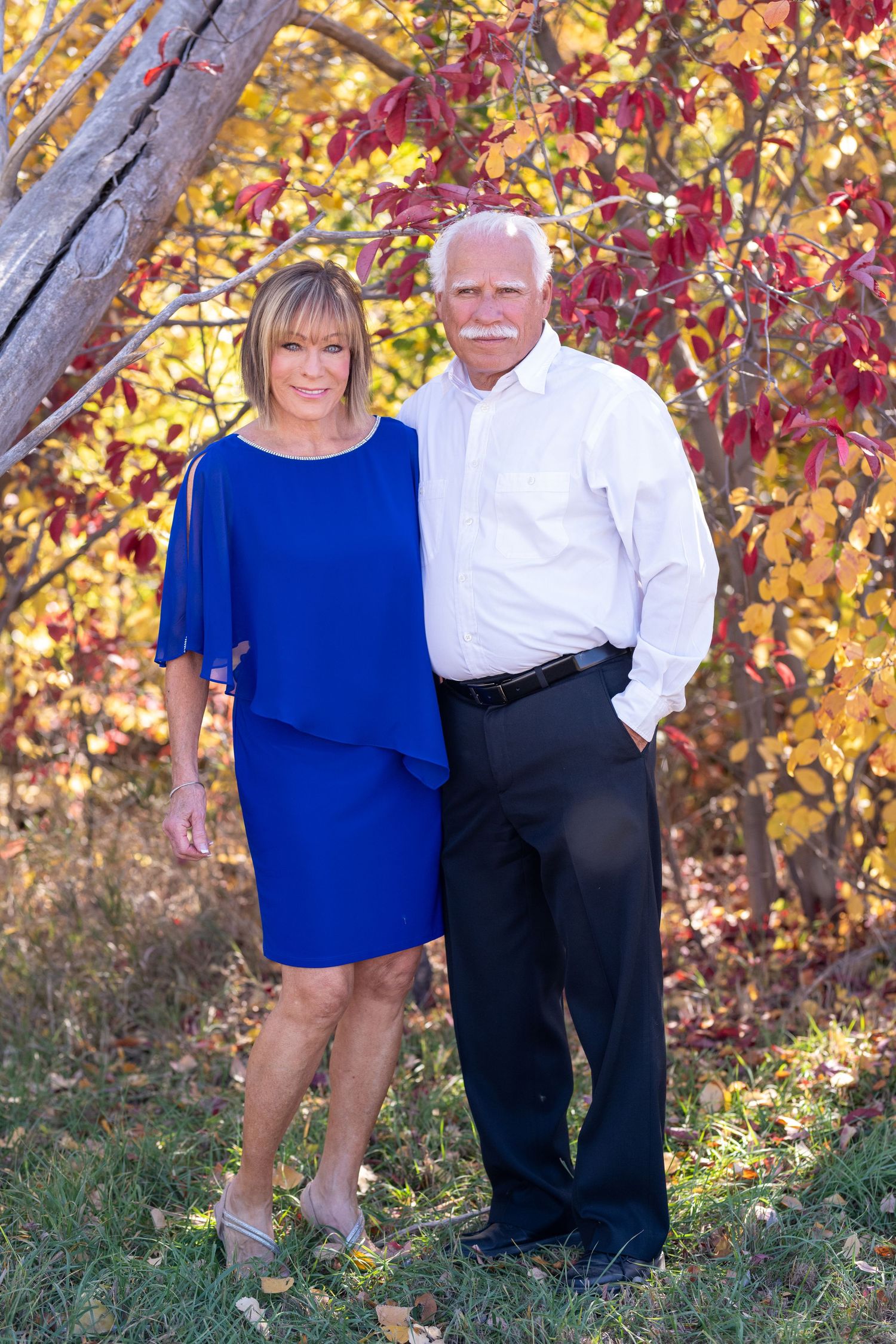 Couple poses together outdoors against autumn foliage background with woman in blue dress and man in white shirt.