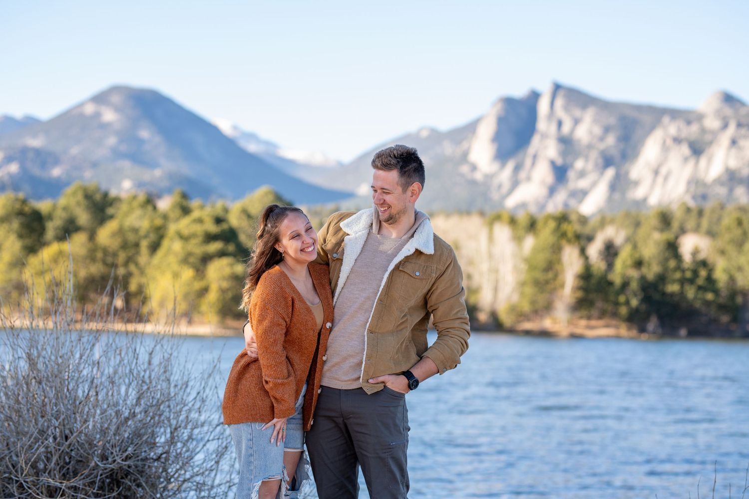 A couple smiles at each other while standing by a lake with rocky mountain peaks in the background.