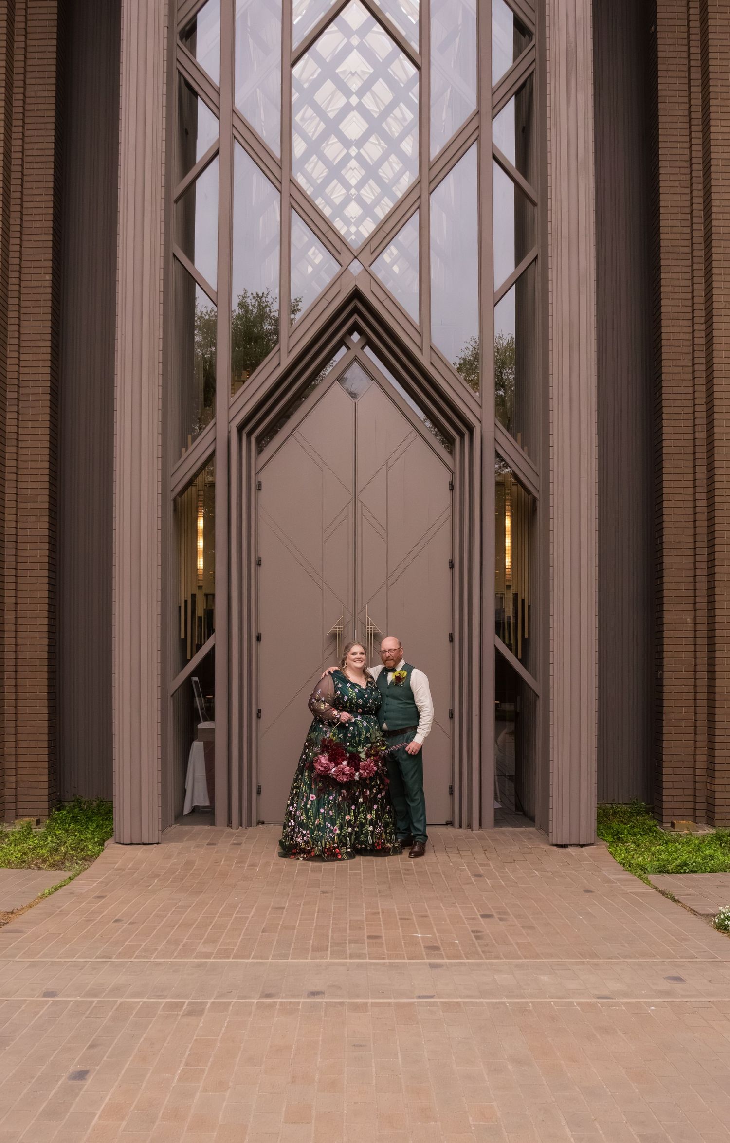 Couple in formal attire posing before grand geometric church doors with tall architectural windows.