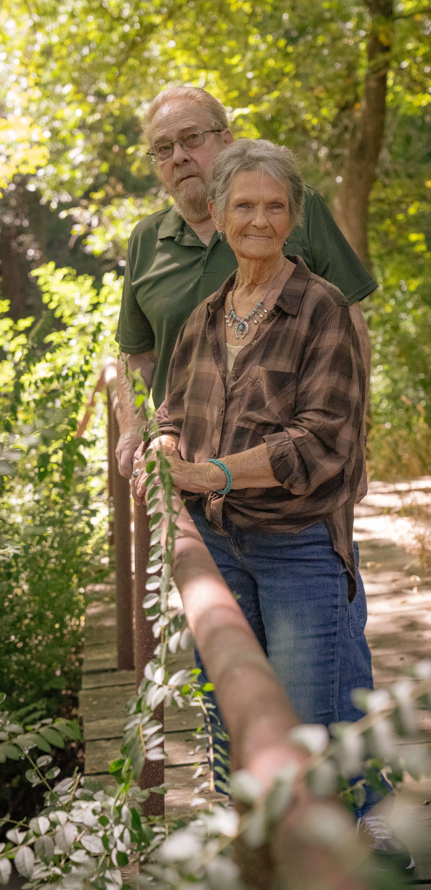 Two people standing close together on a sunlit forest path surrounded by greenery.