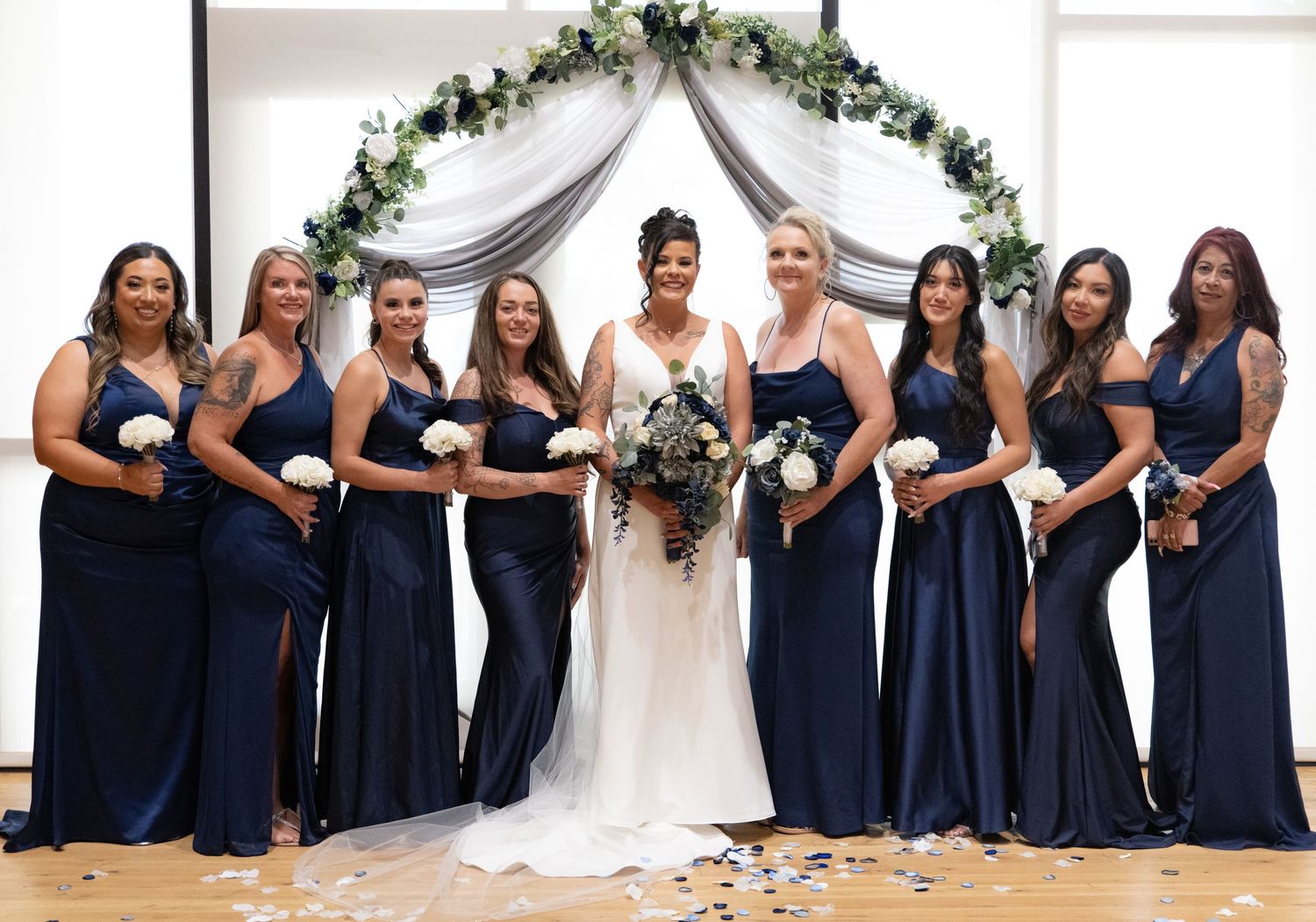 Wedding party in matching navy blue bridesmaid dresses posed under floral arch with scattered petals.