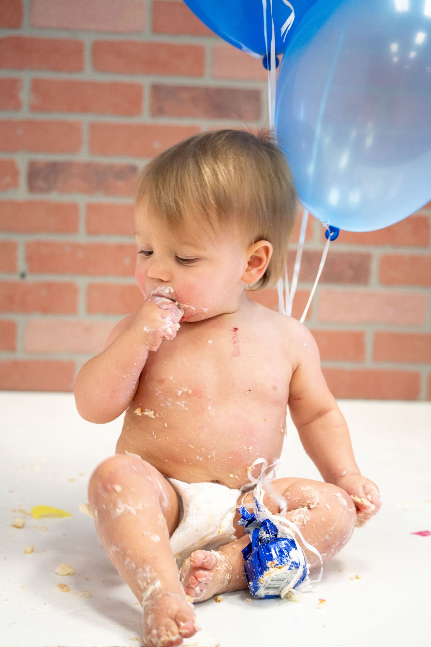 Baby in diaper eating cake with blue balloons and brick wall background.