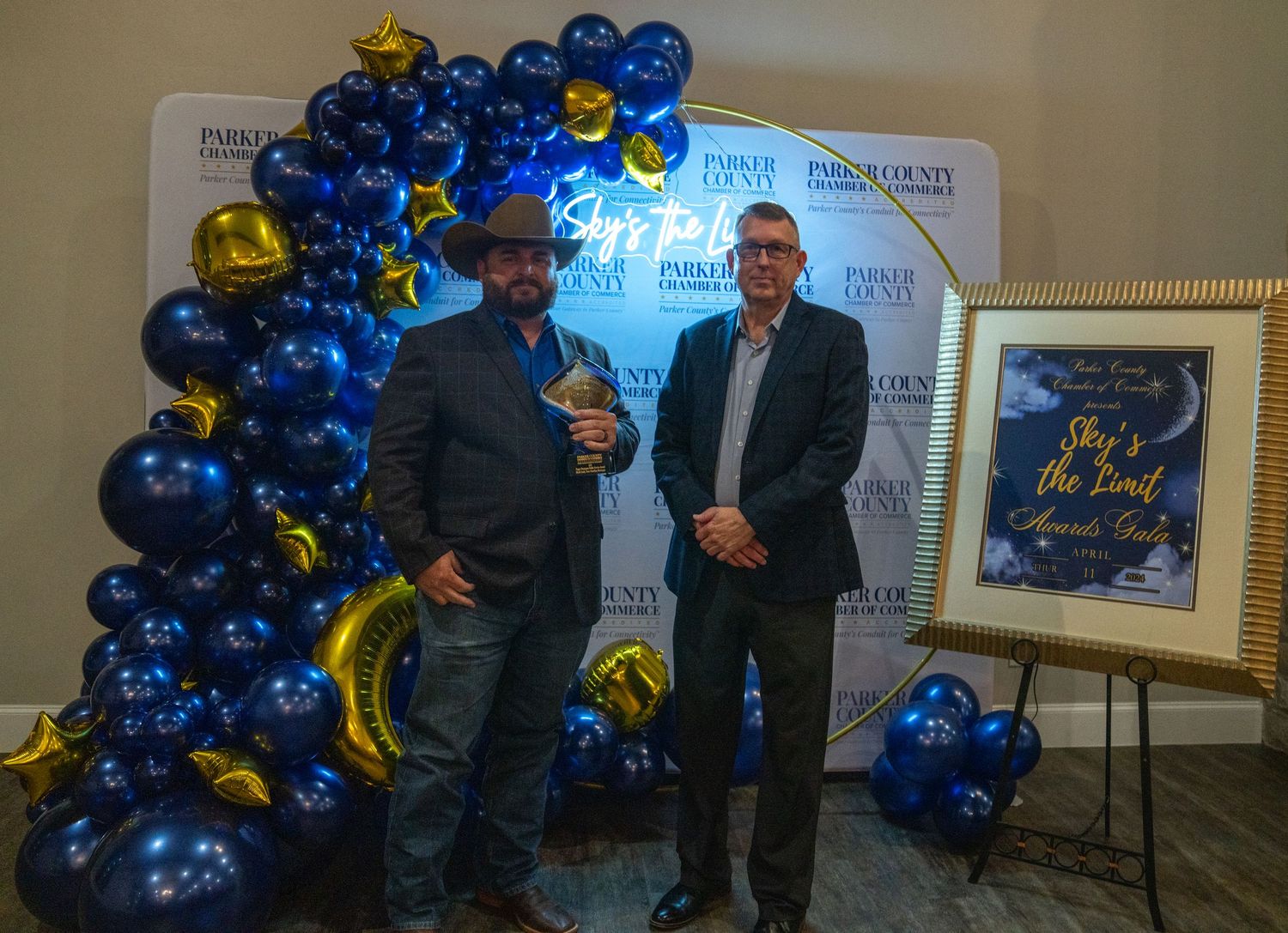 Two attendees stand together at an awards ceremony with blue and gold balloon decorations.