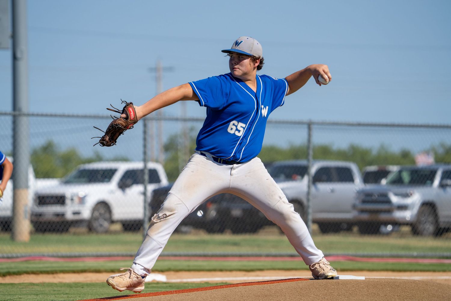 Baseball pitcher in blue jersey winds up for throw on baseball field with cars visible in background.